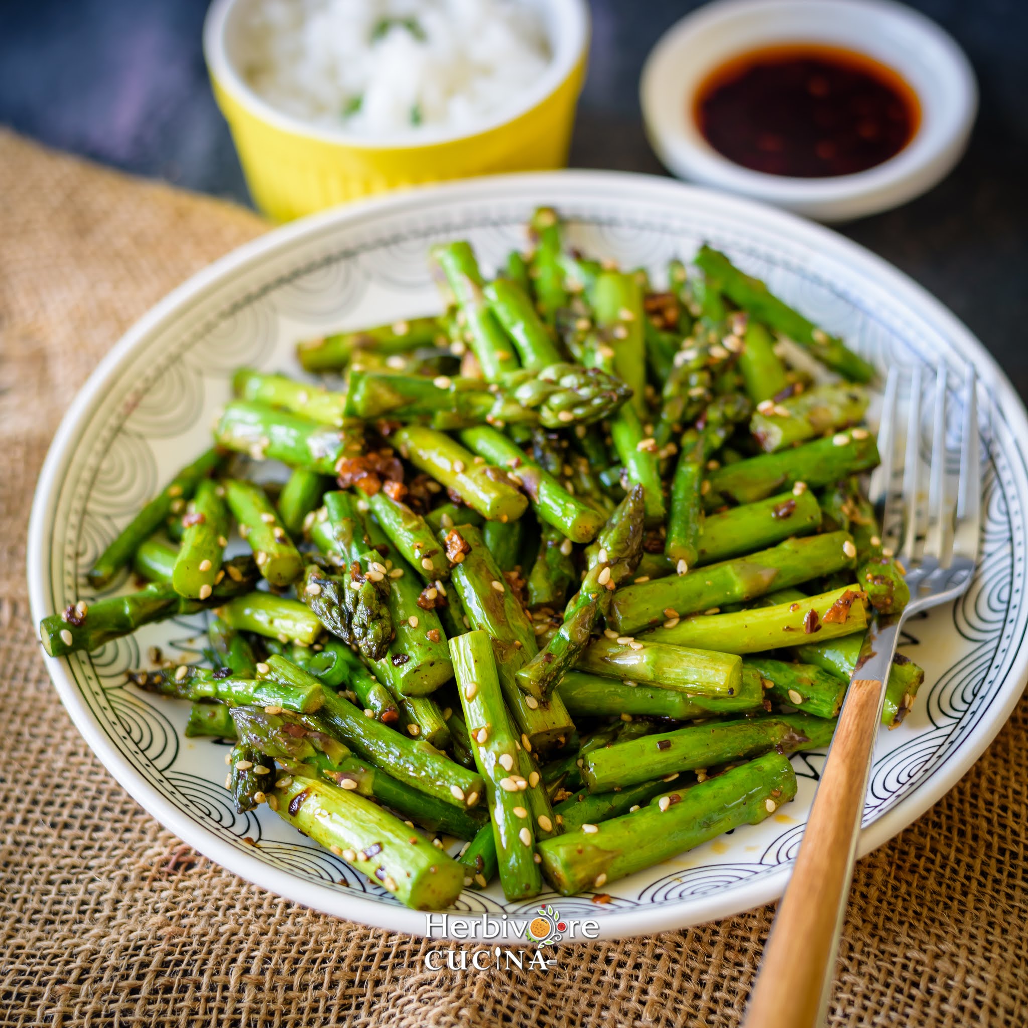 Stir Fried Sesame asparagus in a white plate with some chili oil on top with a bowl of rice on the side. 