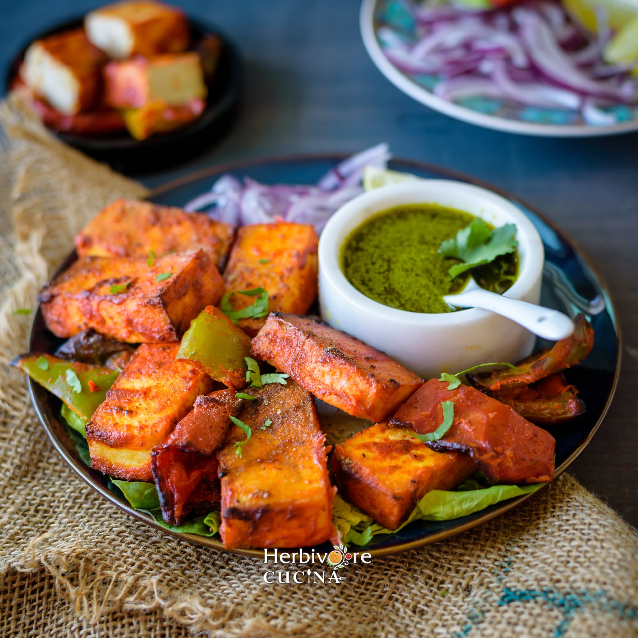 Slices of tofu tikka arranged on a blue plate with green cilantro chutney and sliced onions around it.