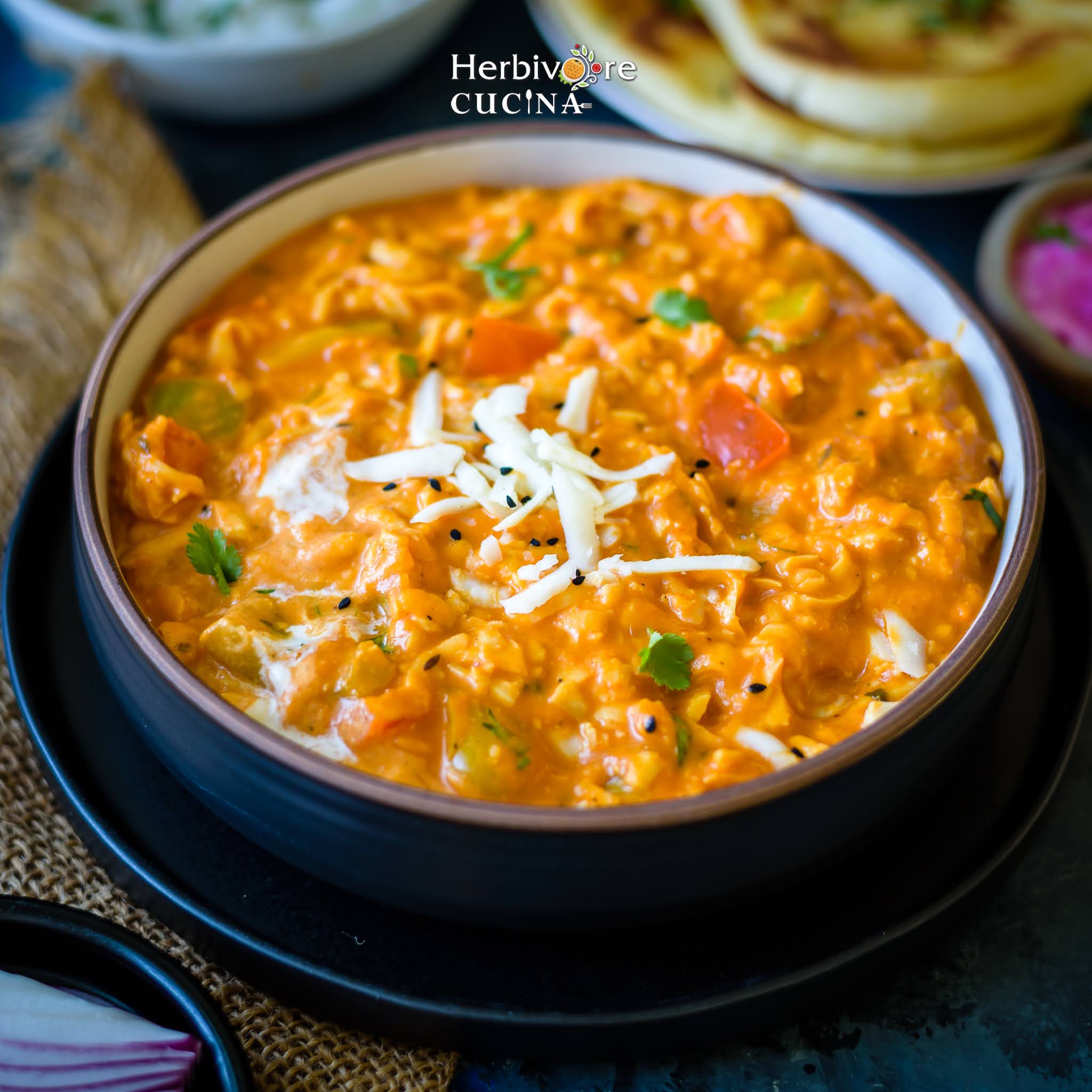 A brown bowl of shredded paneer curry, Akbari Paneer topped with shredded paneer and served with naan and rice in a black plate. 