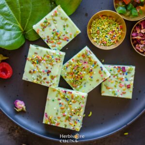 A black plate filled with paan flavored fudge topped with colored fennel, rose petals and sprinkles on a background with betel leaves, fennel and rose around it.