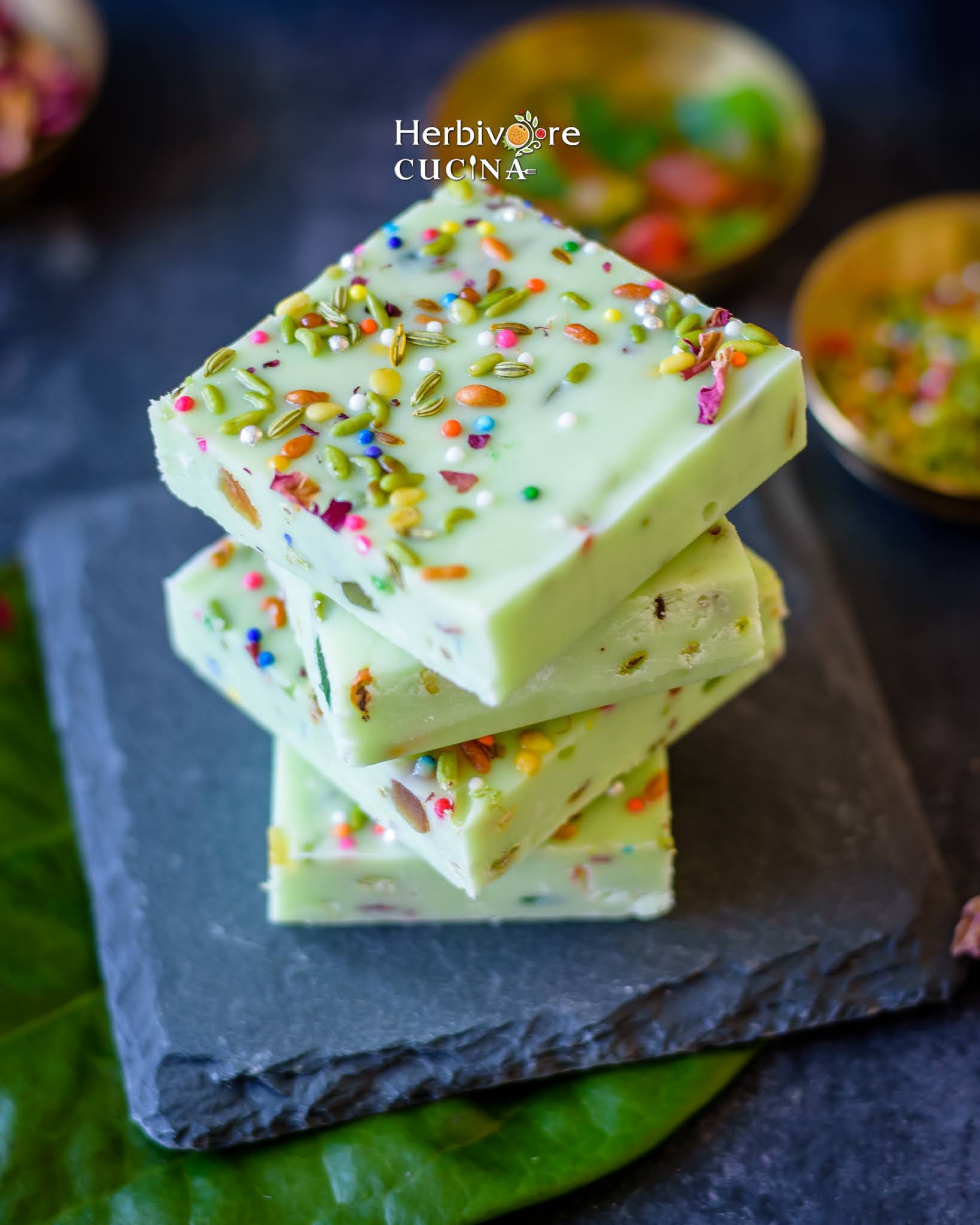 Four pieces of paan fudge on a slate board with the toppings on the side on a dark background. 