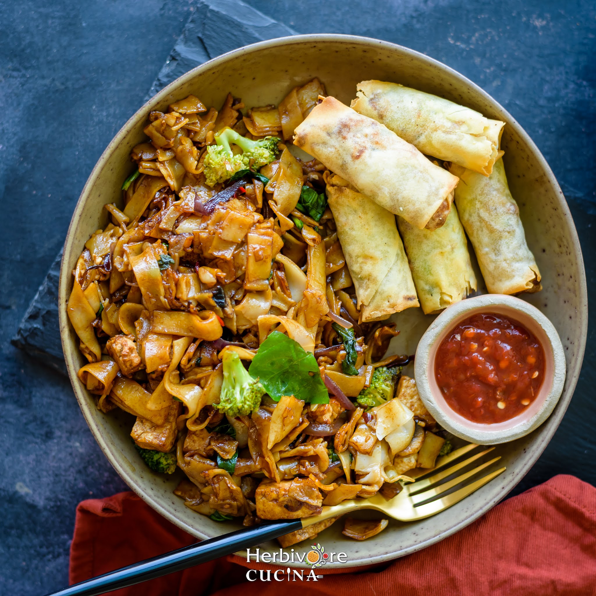 Drunken Noodles in a green plate with egg rolls and hot sauce served with it and a gold black fork beside it on a slate platter. 