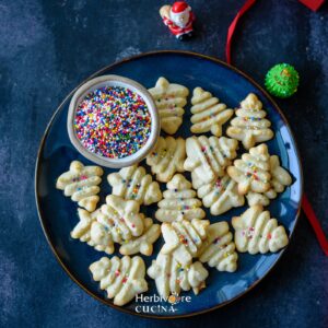 Christmas tree shaped spritz cookies on a plate with sprinkles in a small bowl.