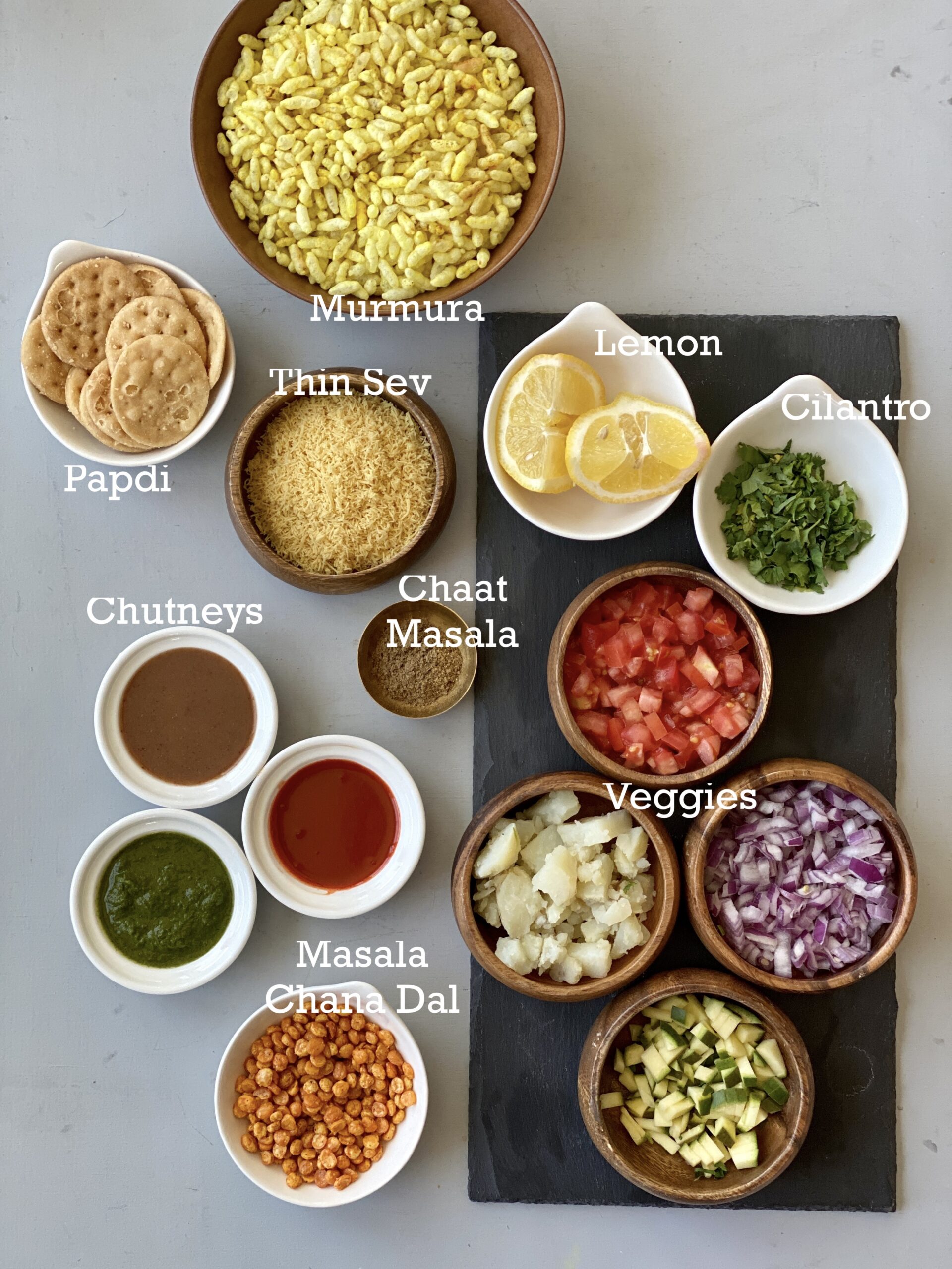 Ingredients to make bhel puri in small bowls on a gray surface.