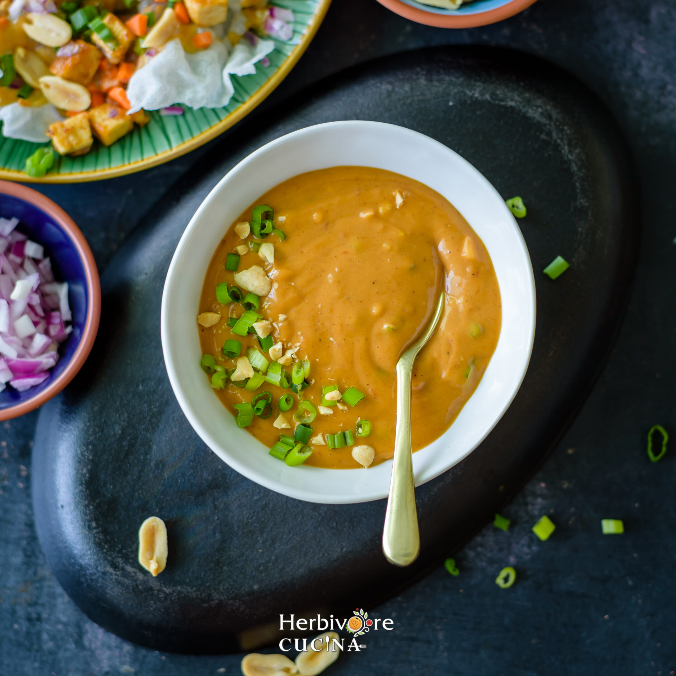 Top view of Peanut Sauce in a white bowl against a dark background with chopped onions and Spring Roll Nachos on the side. 