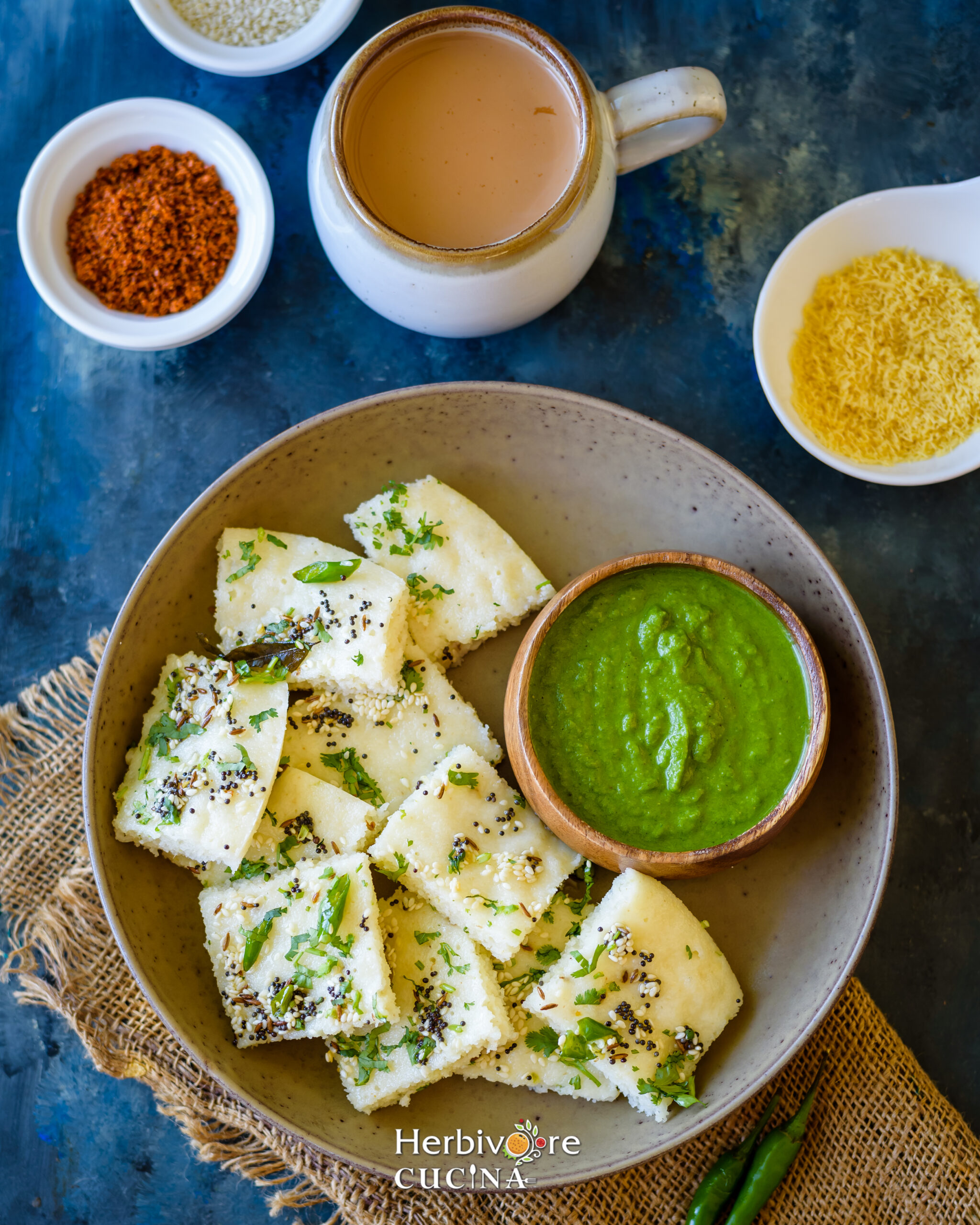 Rava Dhokla served in a brown plate with chai and chutney around it on a blue black background. 
