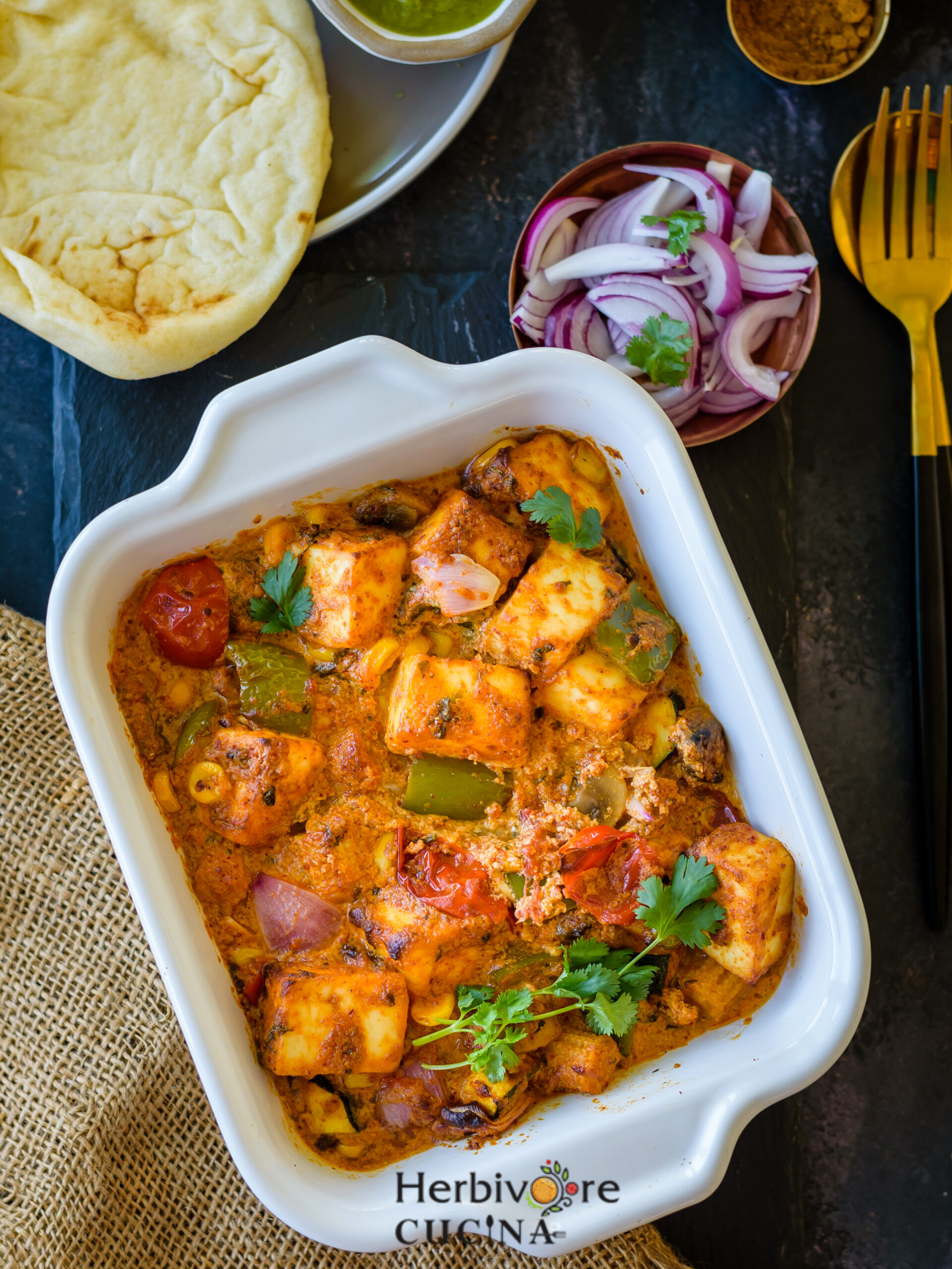 malai paneer, a mix of marinated paneer, peppers and onions baked in the sire fryer served in a white rectangle tray on a black surface. 