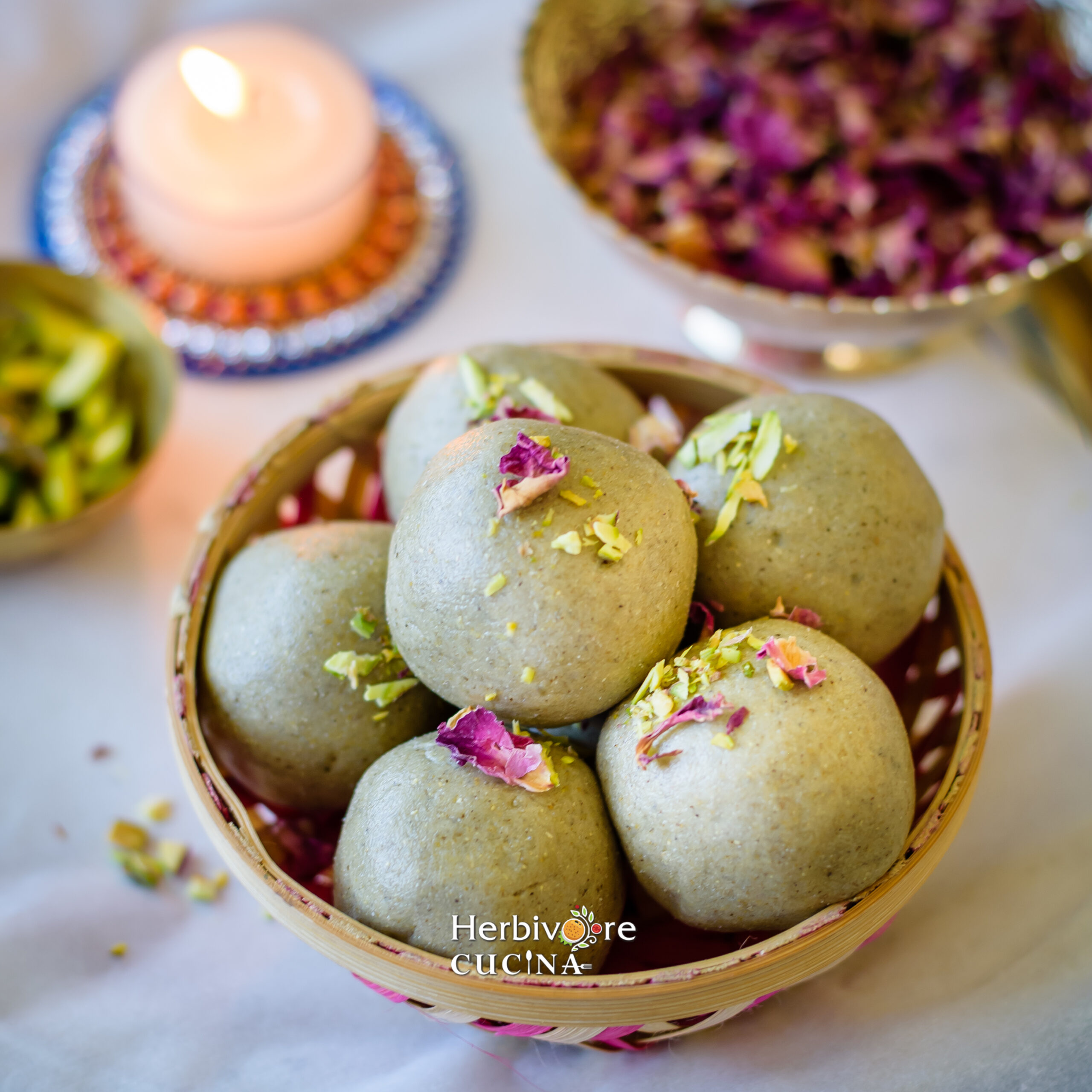 Kuler Ladoos in basket topped with pistachio and rose petals with rose petals and a candle in the background on a white board. 