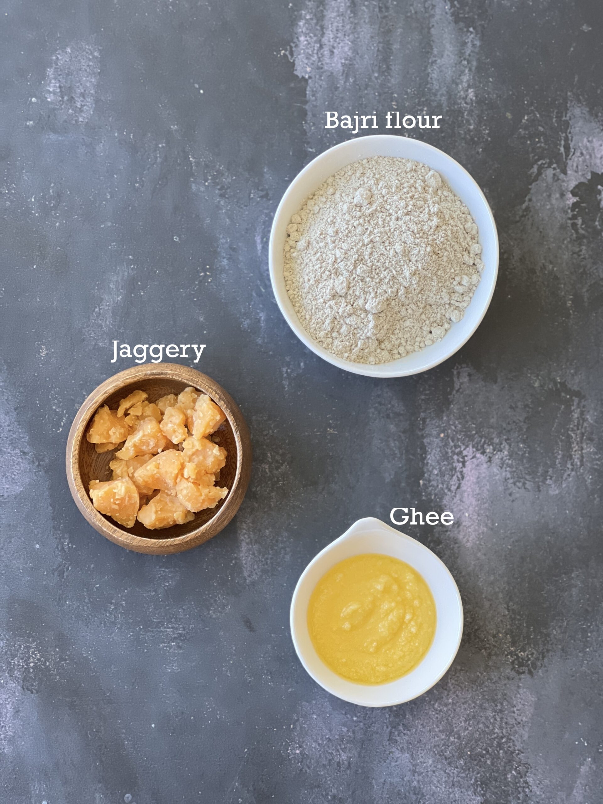 Ingredients for Kuler Ladoo; jaggery, bajri flour and ghee in bowls on a black board. 