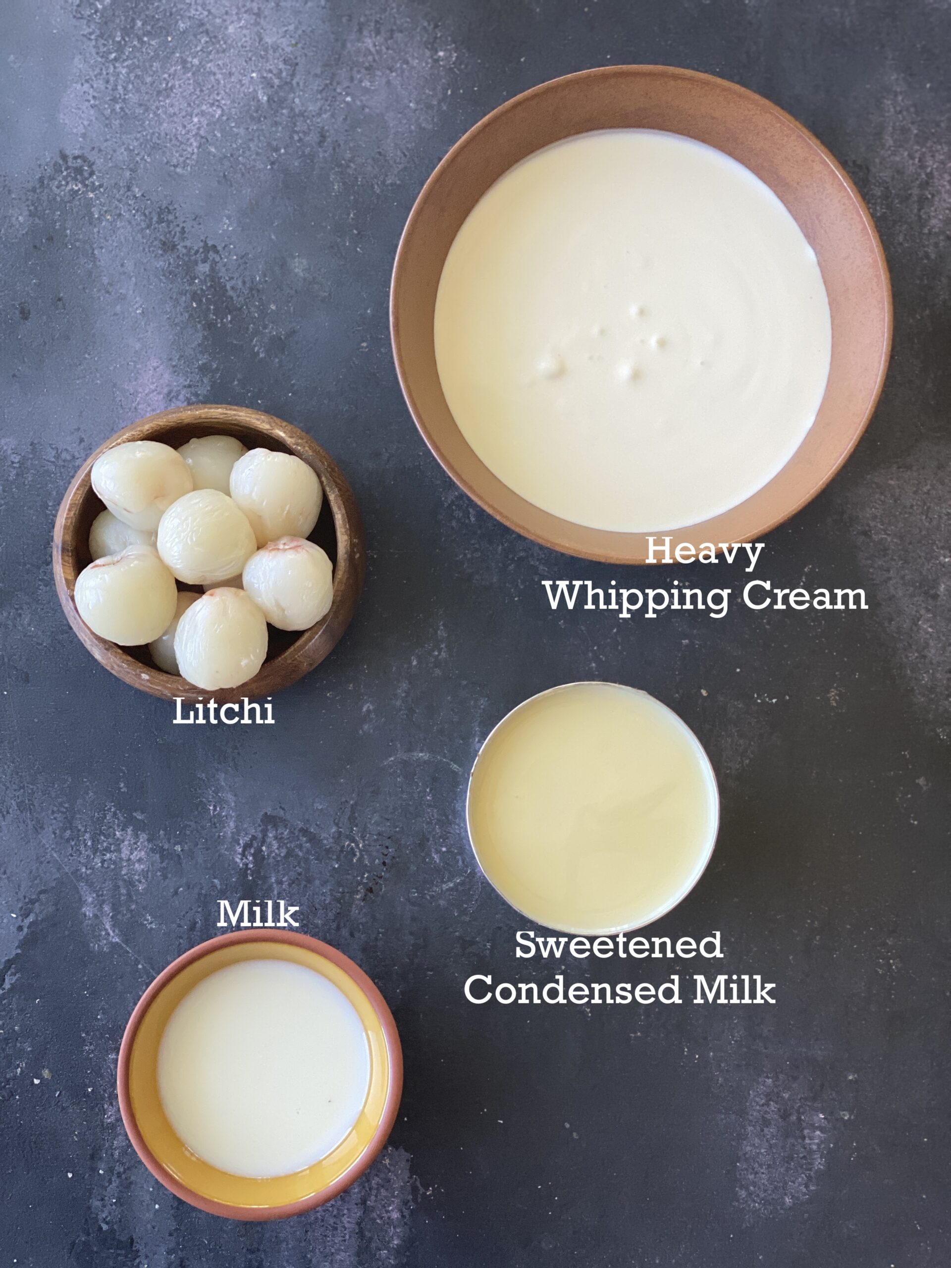 Ingredients for litchi ice cream in small bowls; lychee fruit, condensed milk, milk and whipping cream on a black surface. 