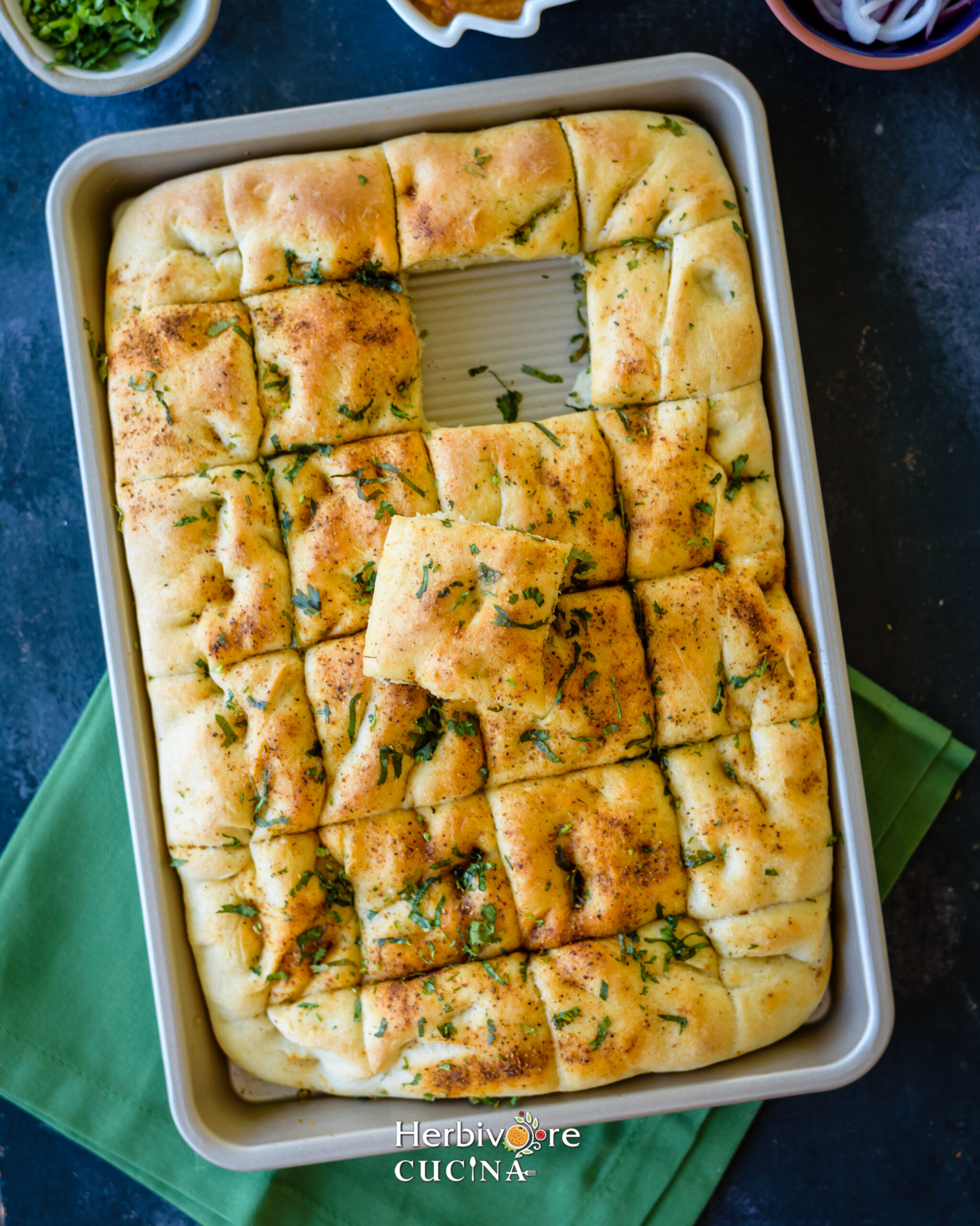 A half baking tray with Focaccia Bites and one piece removed and placed on top on a green napkin. 