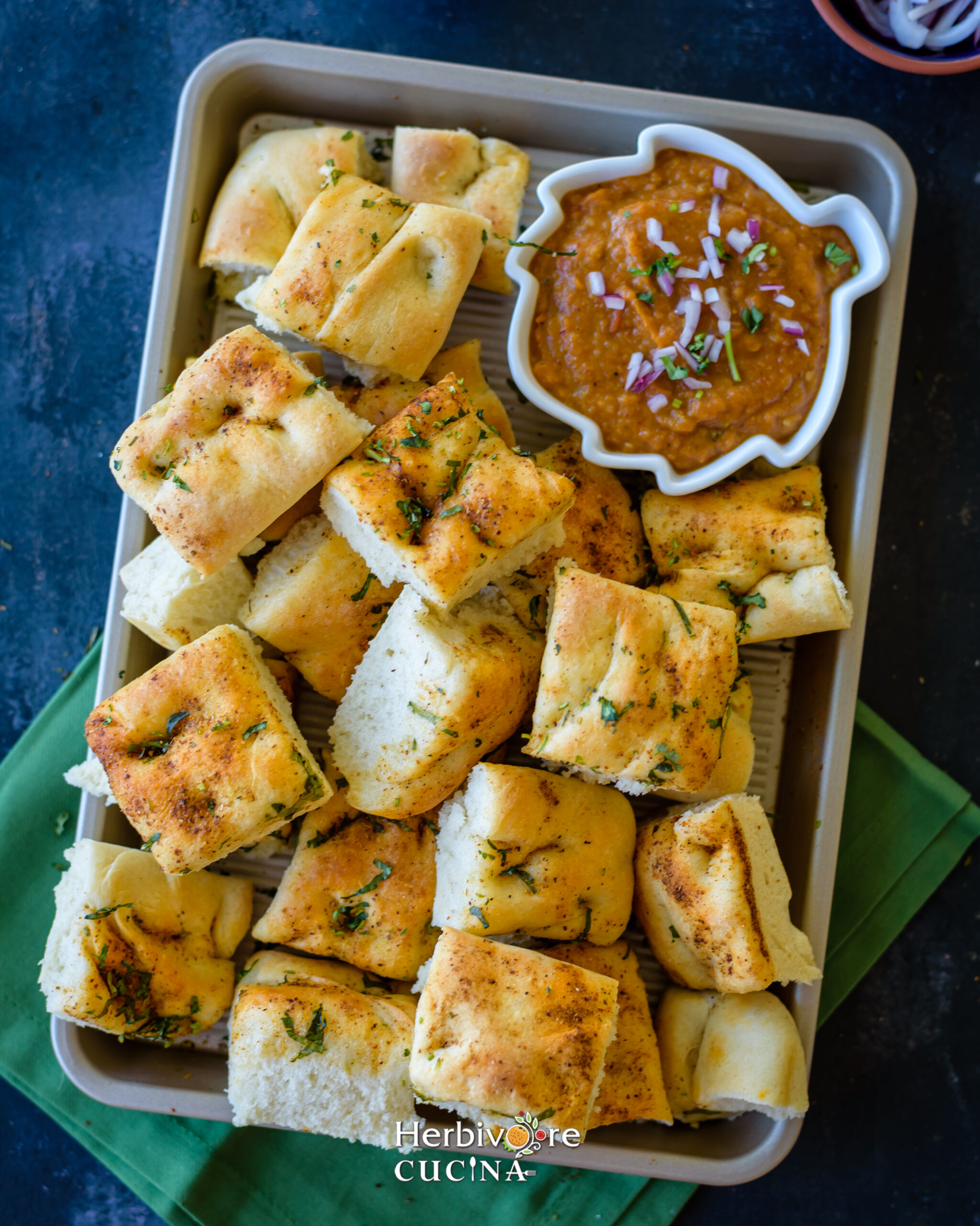 Bites of Pav Bhaji Focaccia in tray with some bhaji in a bowl on the side topped with diced onions. 