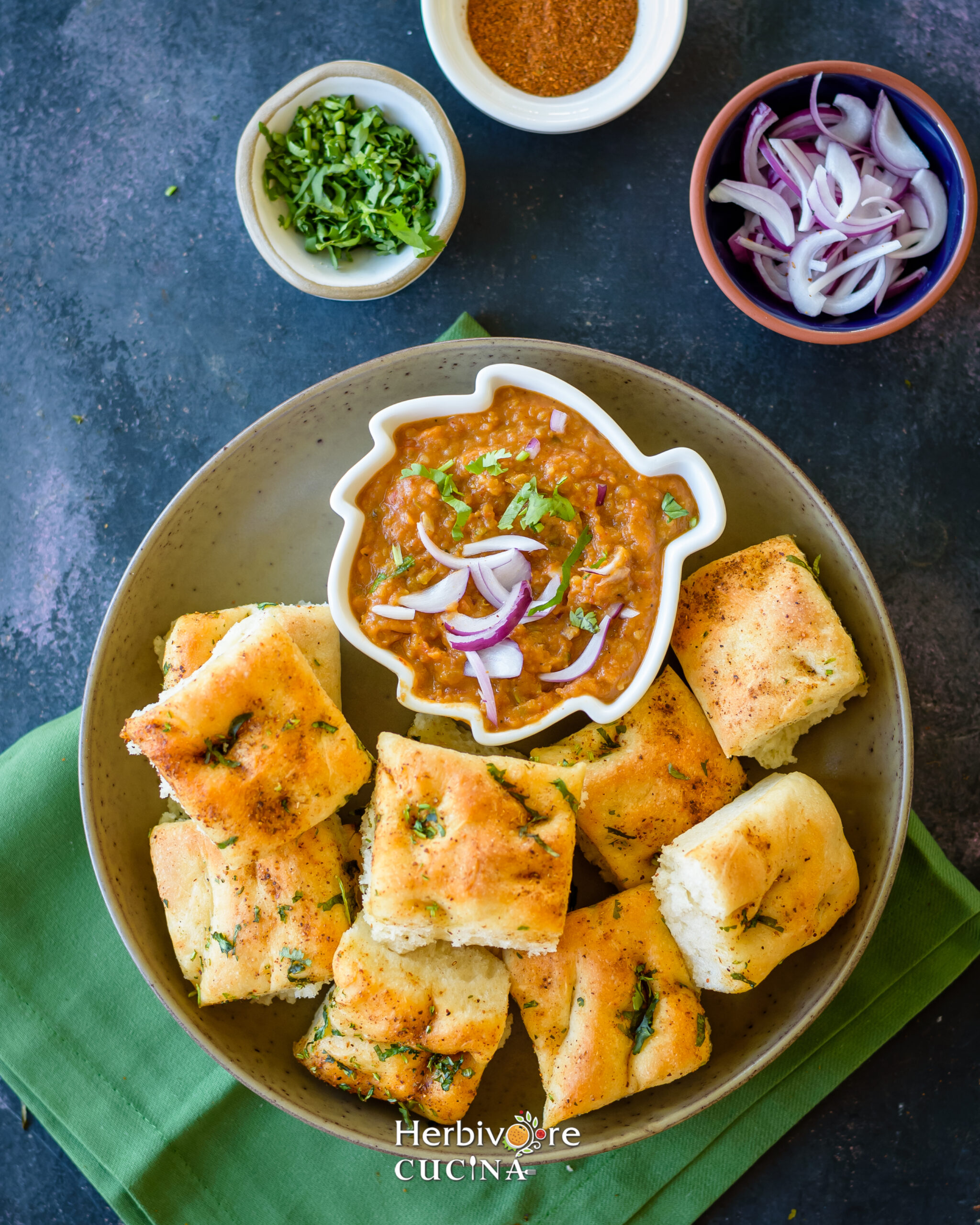 A brown plate filled with bites of baked focaccia bread and served with a bowl of pav bhaji topped with onions on a black surface. 