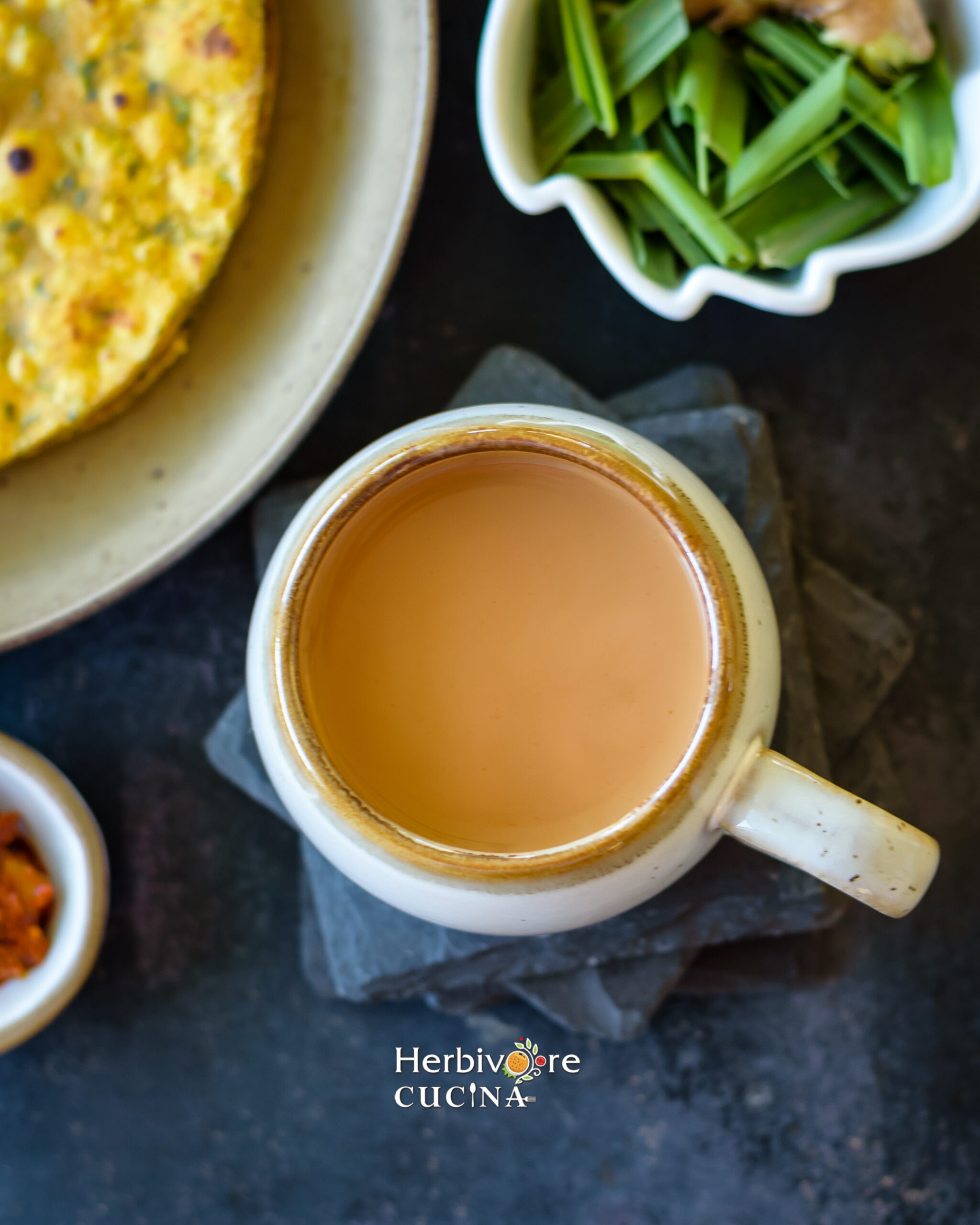 Top view of masala chai in a white cup with some thepla and tea leaves on the side on a slate plate. 