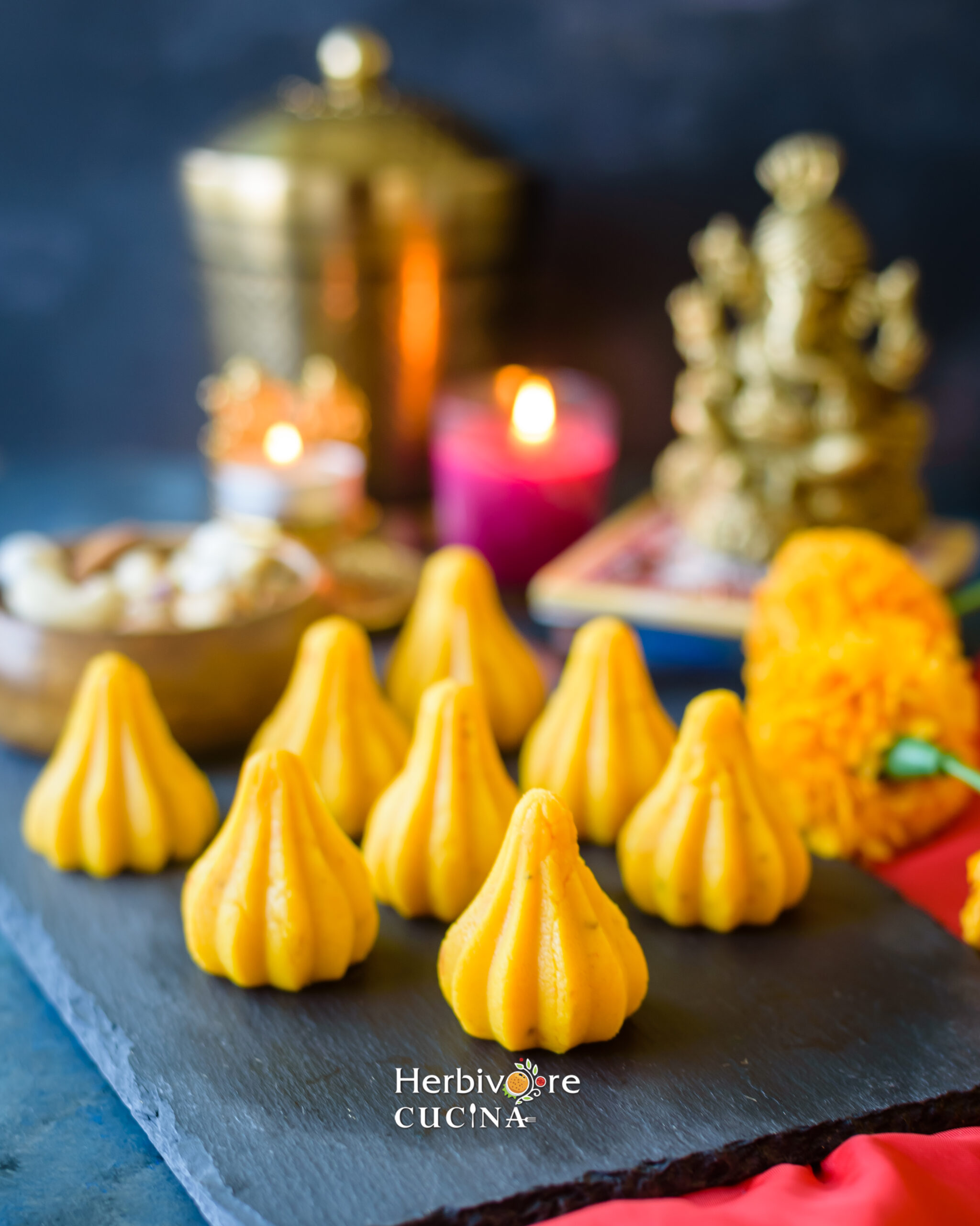 Yellow Kesar malai Modak arranged on a slate platter with an idol of Ganesha, flowers and candles in the background. 