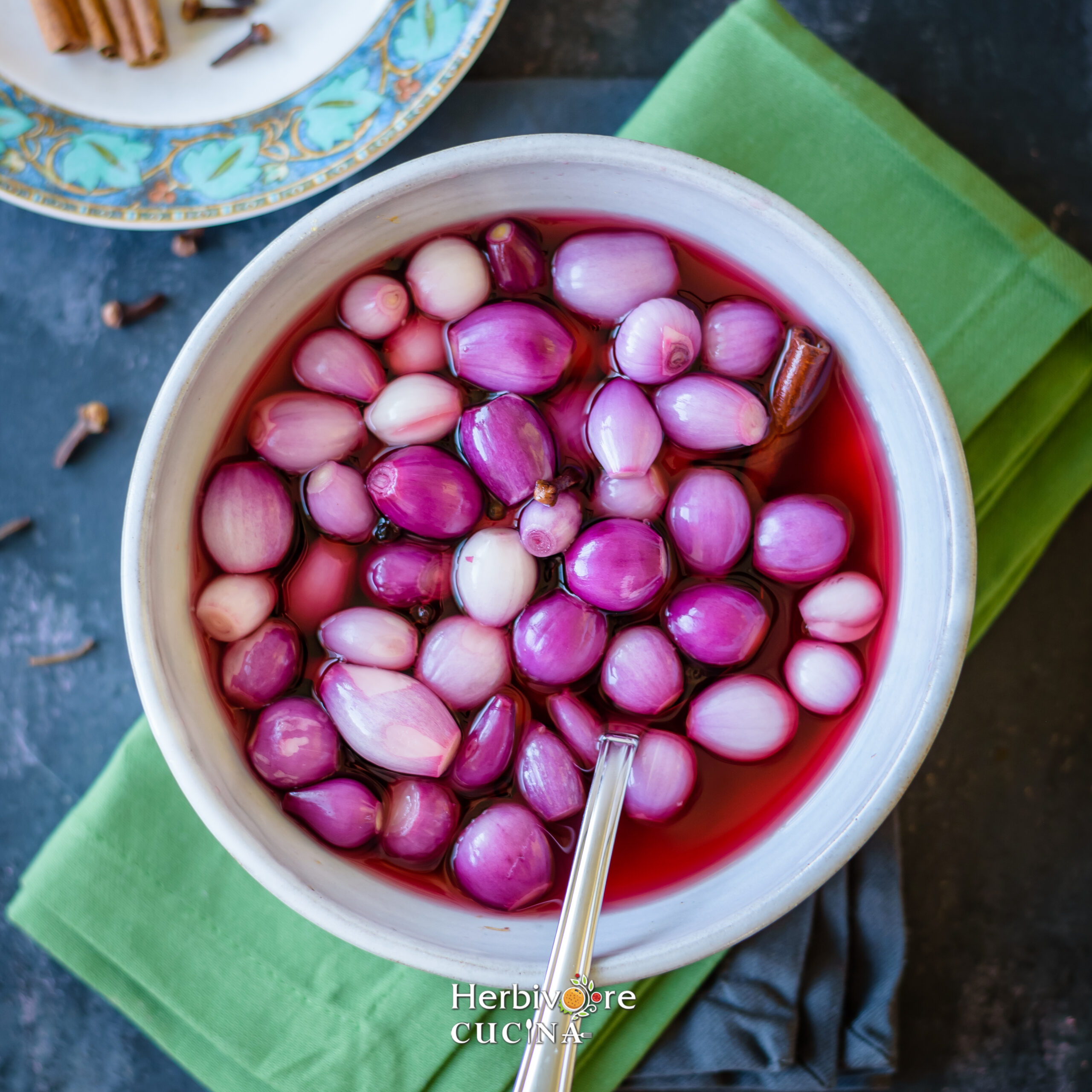 Pickled Onions in beetroot brine in a gray bowl placed on a board with green napkin below it. 