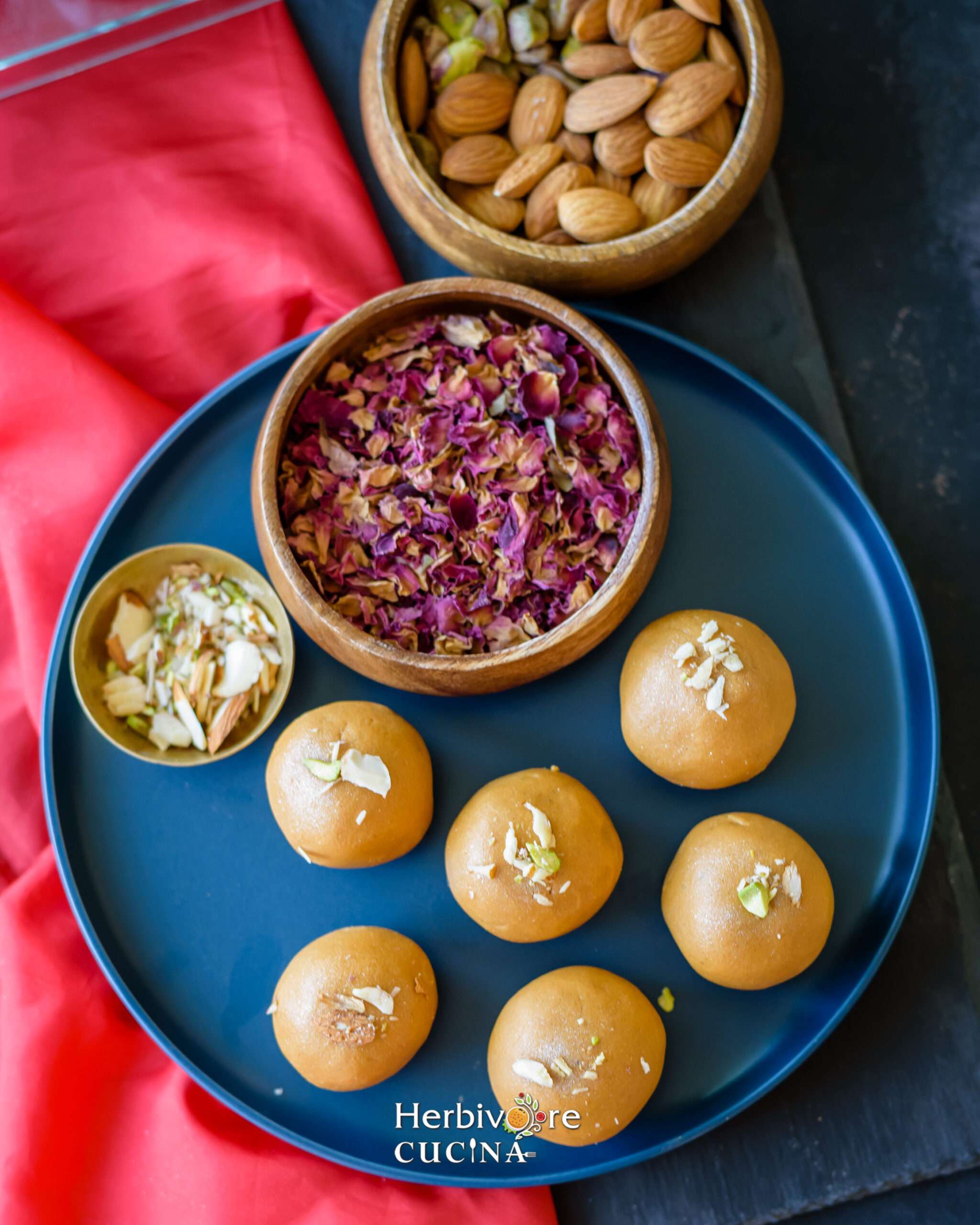 Top view of besan ladoo in a blue plate with rose petals and slivered nuts in small bowls around it. 