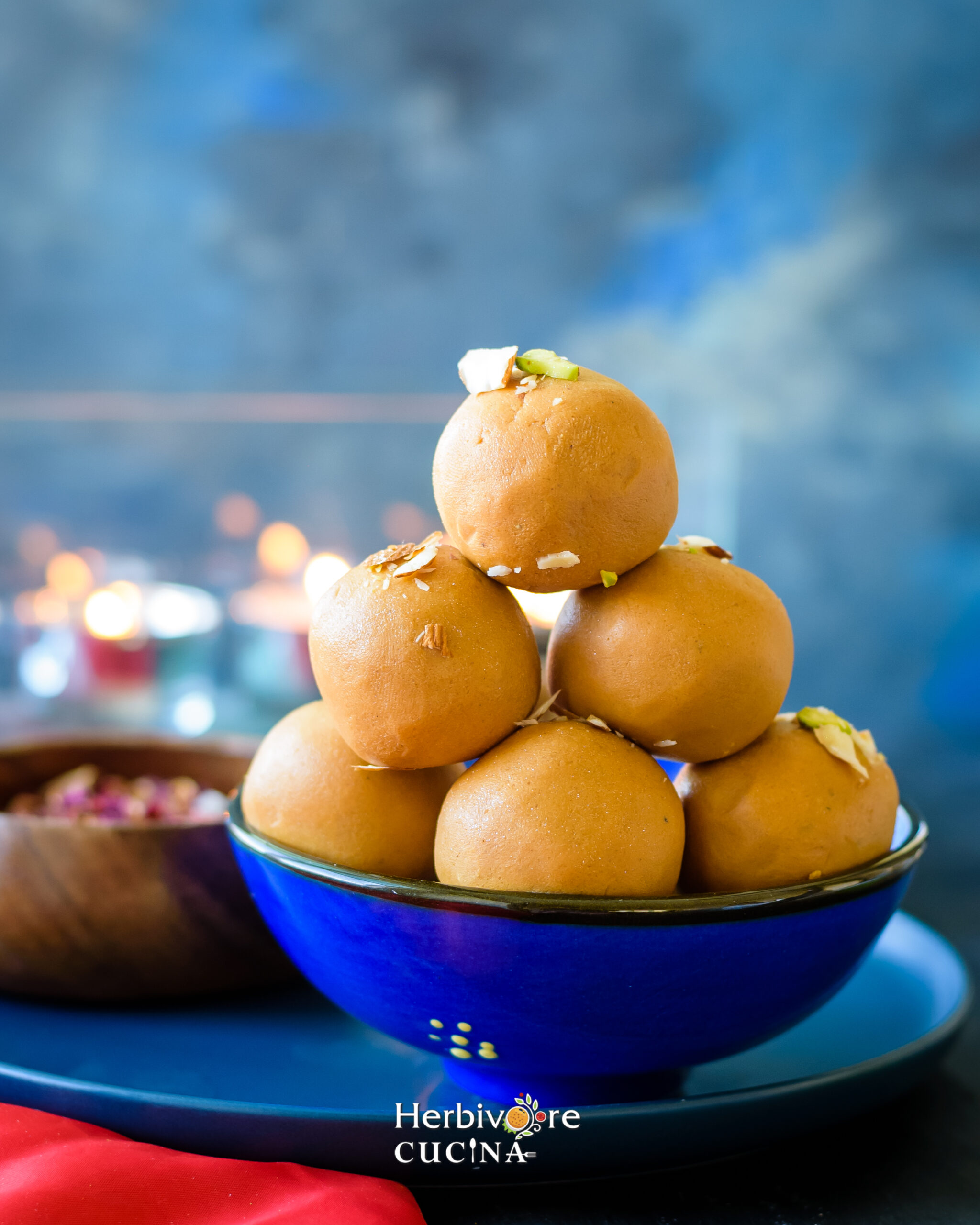 Six Besan Ladoos piled on a blue bowl with some rose petals in the background on a blue plate. 