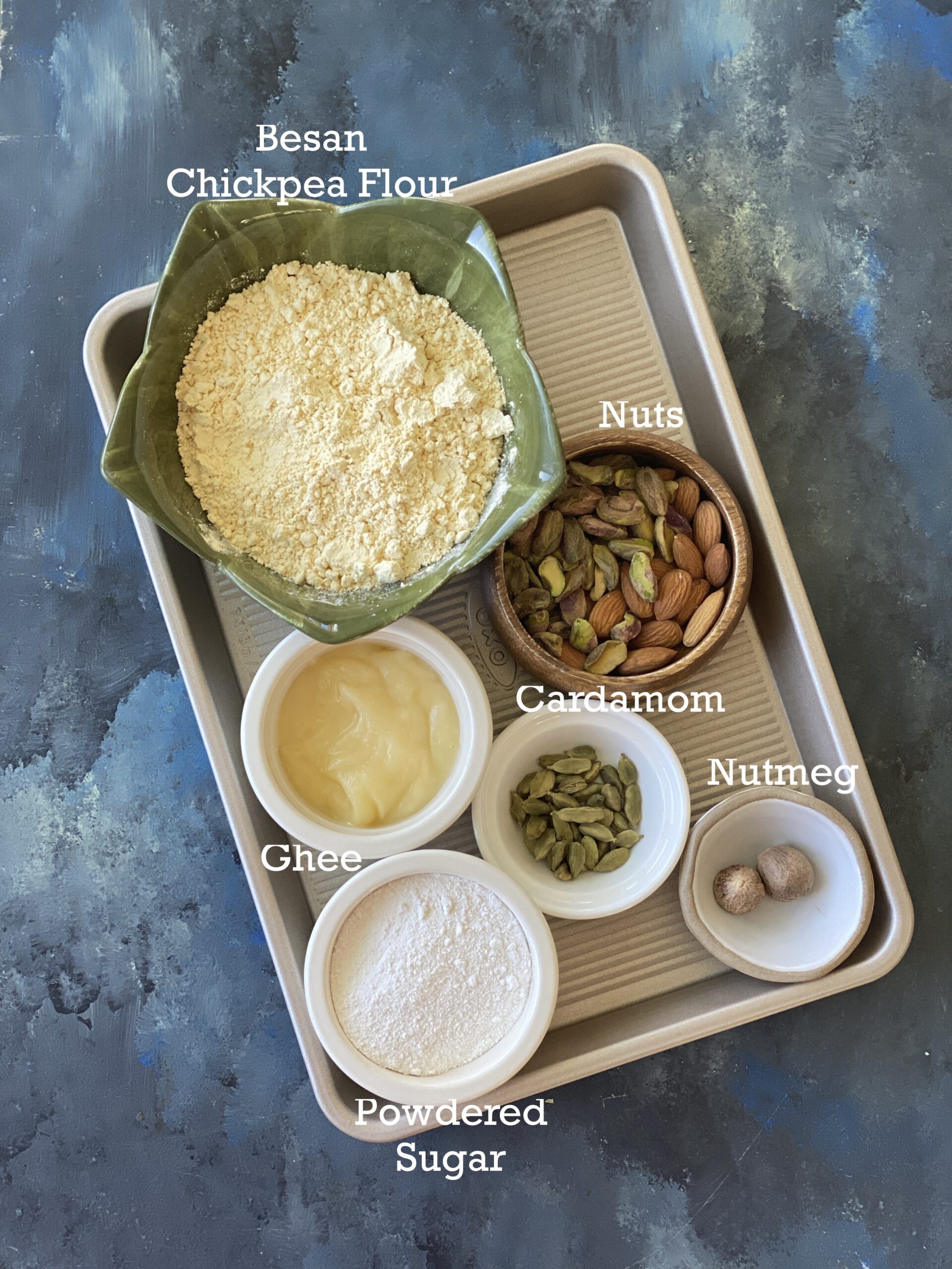 Ingredients for Besan Ladoo; besan, ghee, sugar and add-ins in small bowls on a metal tray. 