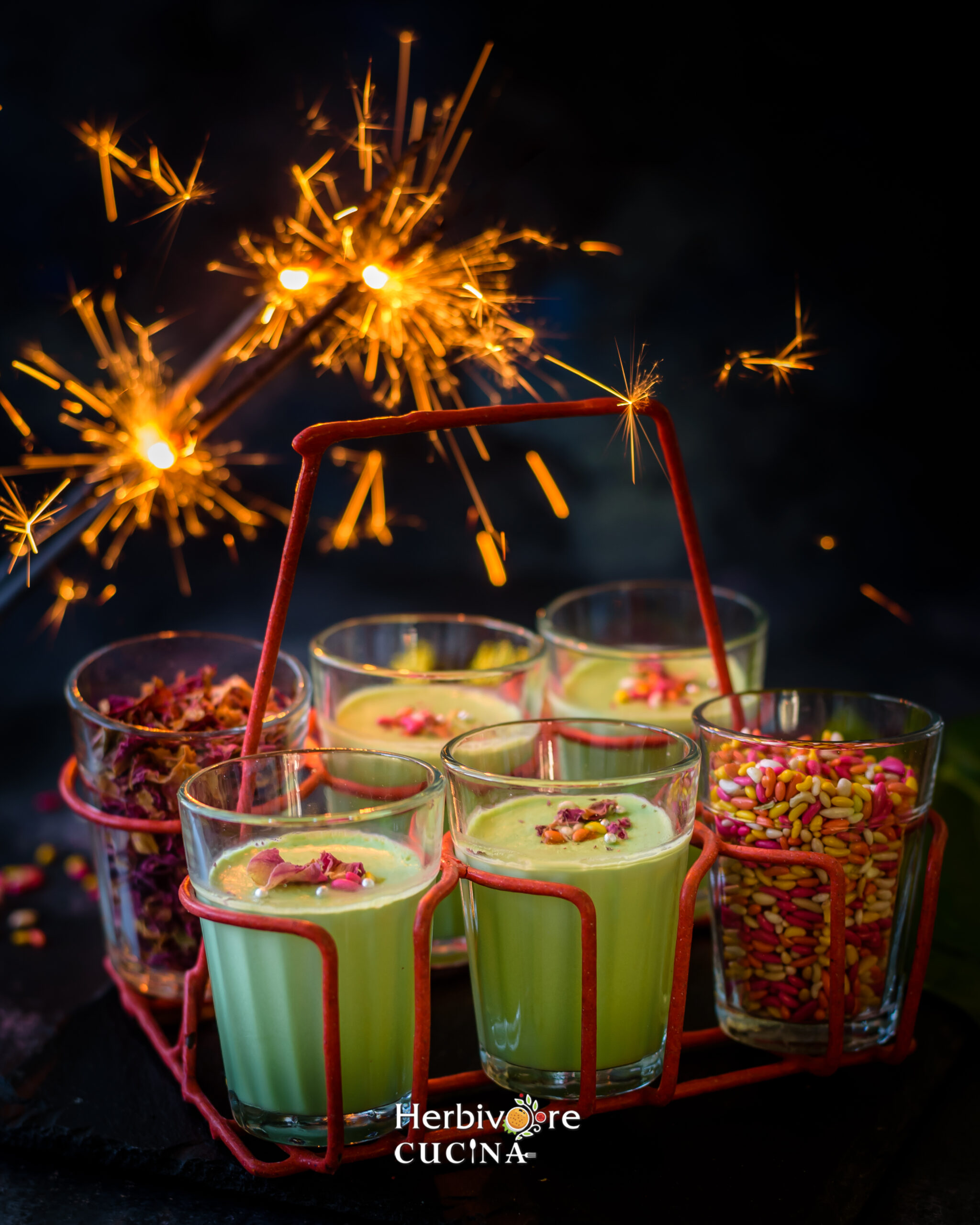 Four bowls of paan panna cotta and two bowls with add-ins against a lighted firework on a dark background. 