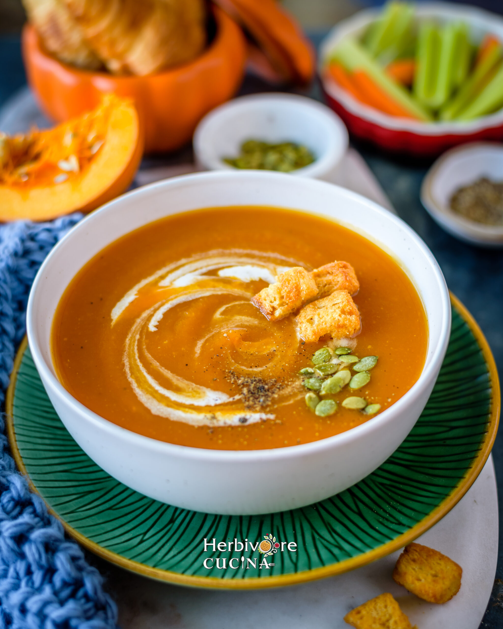 Pumpkin Soup in a white bowl placed on a green plate with pepitas, cream and croutons on top and more vegetables placed behind it. 
