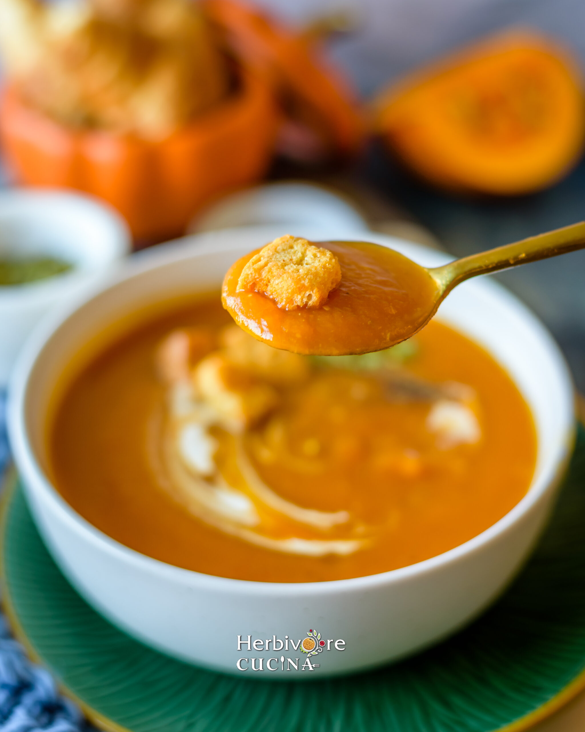 A spoon full of pumpkin soup; help over a bowl of pumpkin soup with more pumpkins in the background. 