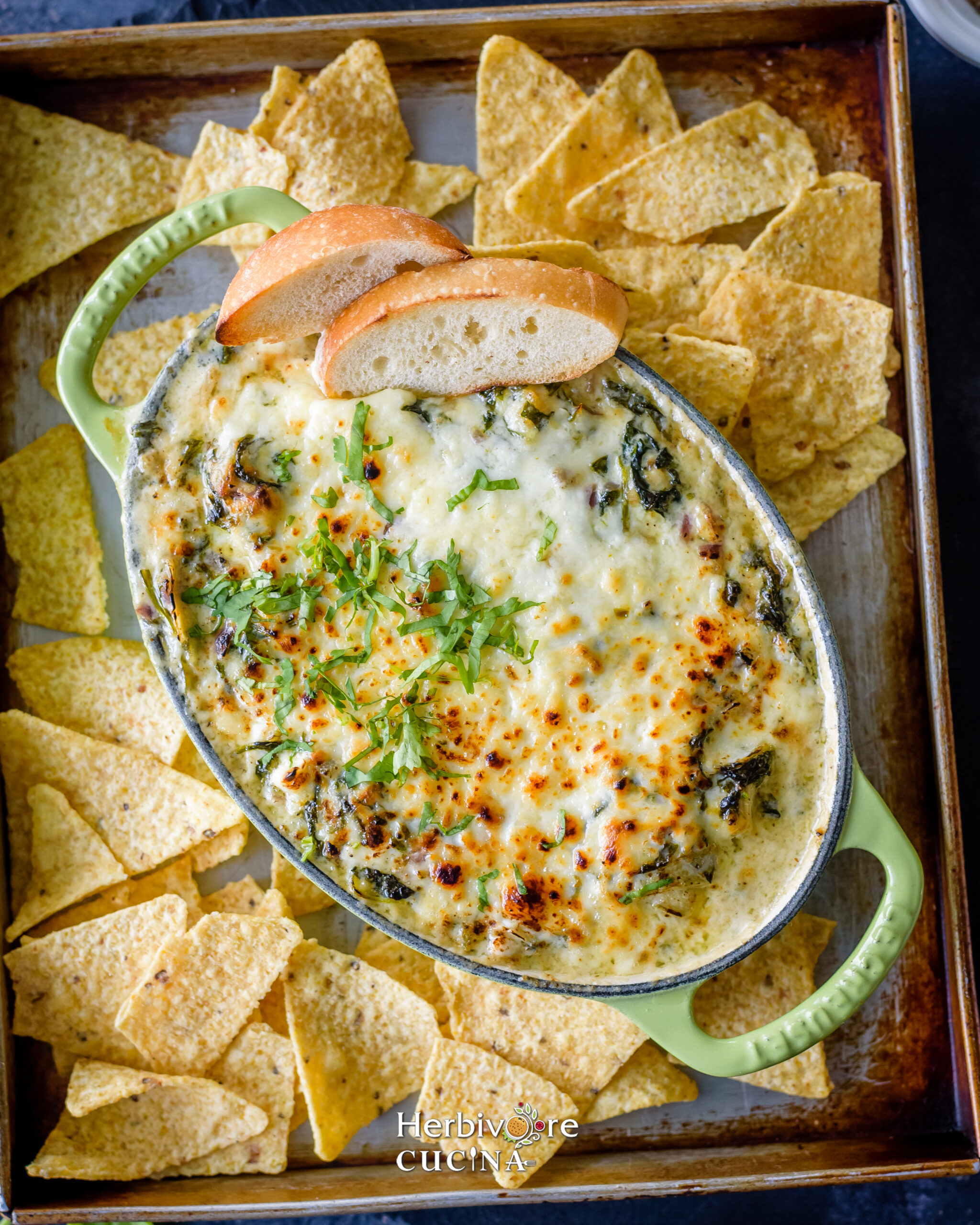 Baked Spinach Artichoke Dip in a green bakeware with chips and bread slices around it in an aluminum platter. 