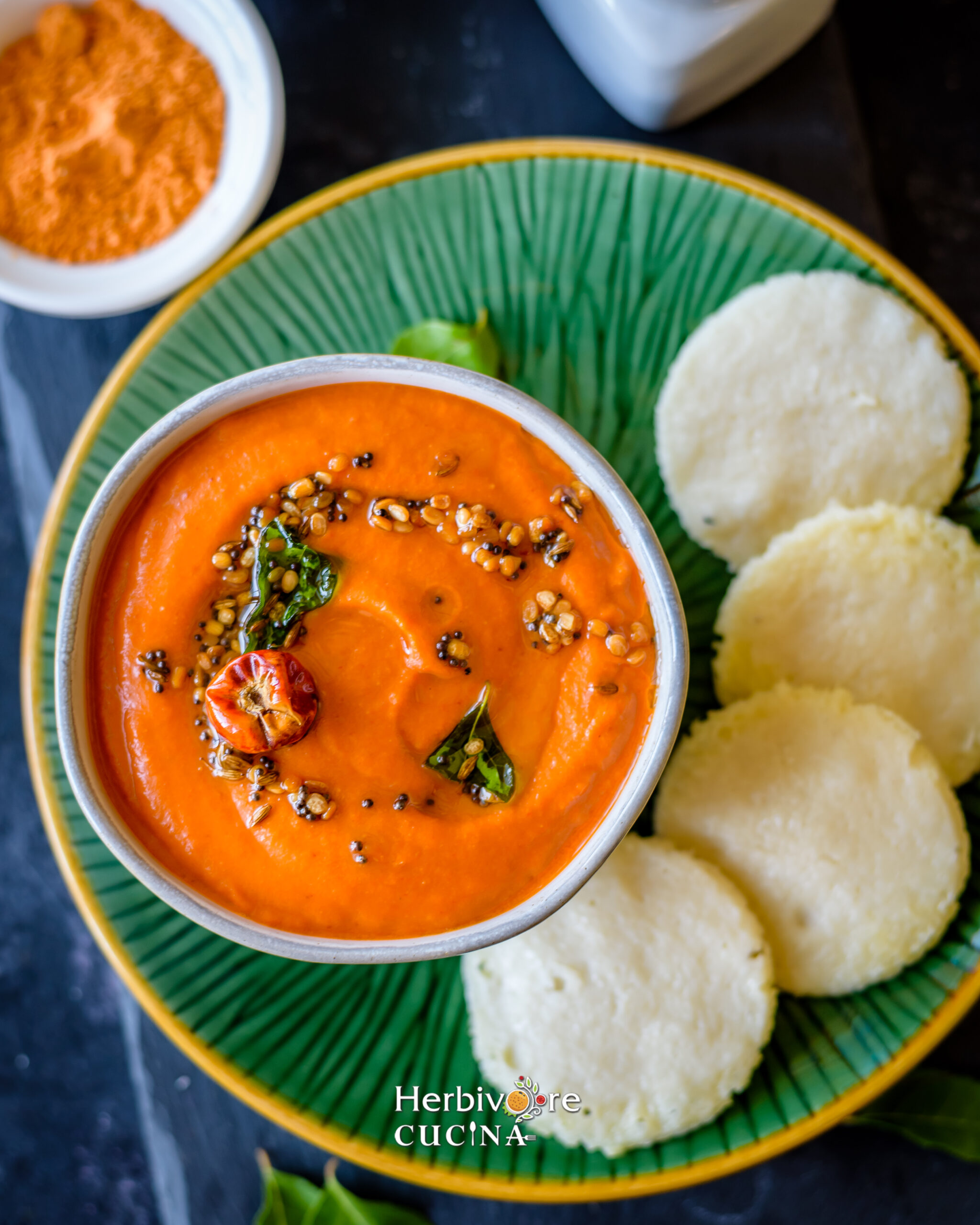 Top view of roasted tomato onion chutney with idli served in a green plate on a dark background.