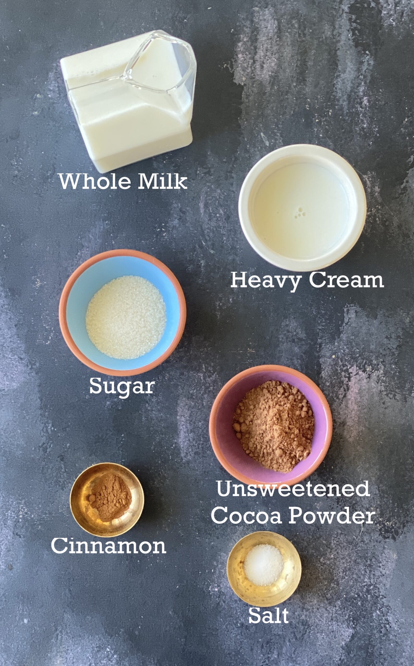 What you need to make Christmas hot cocoa; cocoa powder, sugar, salt, milk and toppings arranged in bowls on a black surface. 