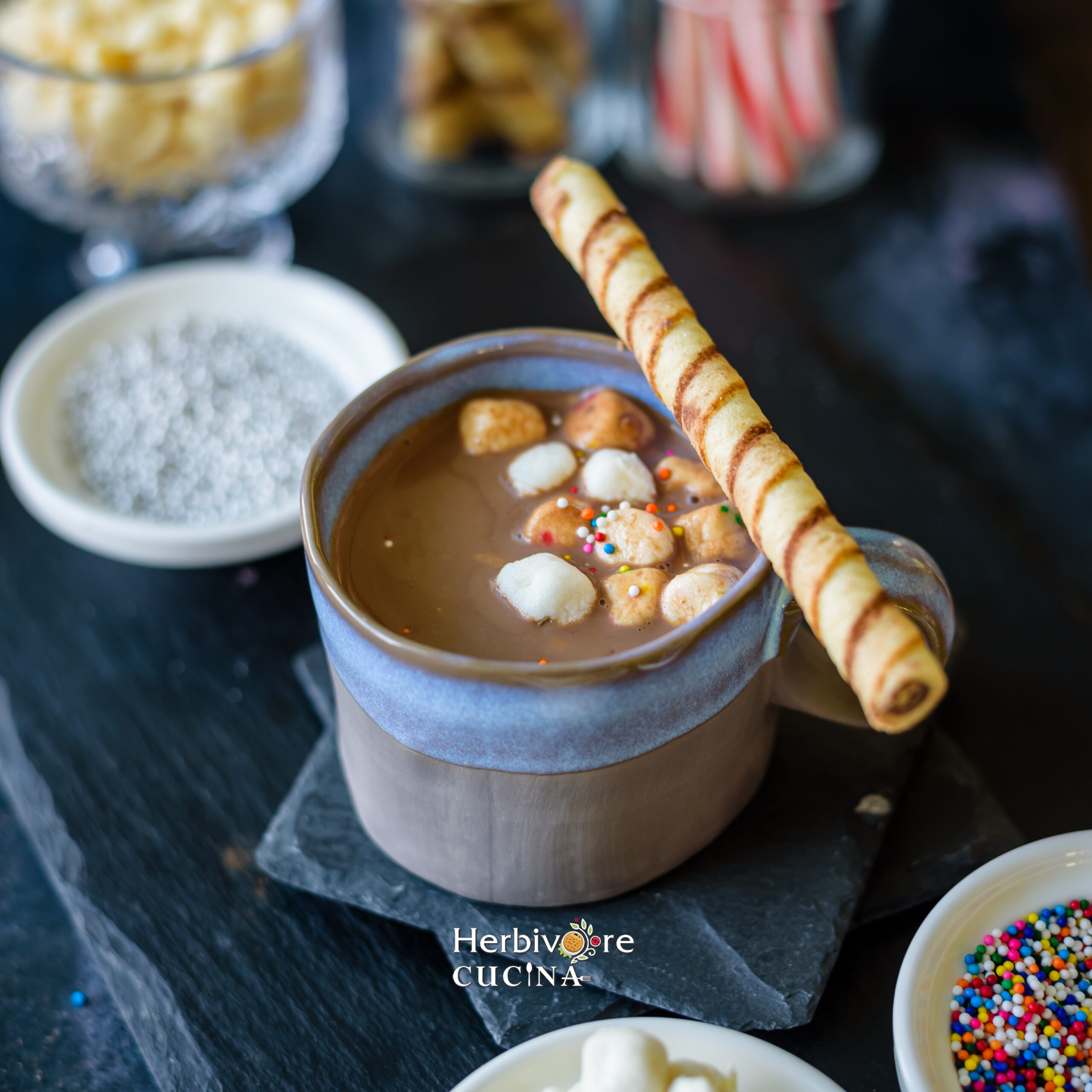 Hot Chocolate served in a blue mug topped with marshmallows, sprinkles and a chocolate stick on he side on a slate platter. 