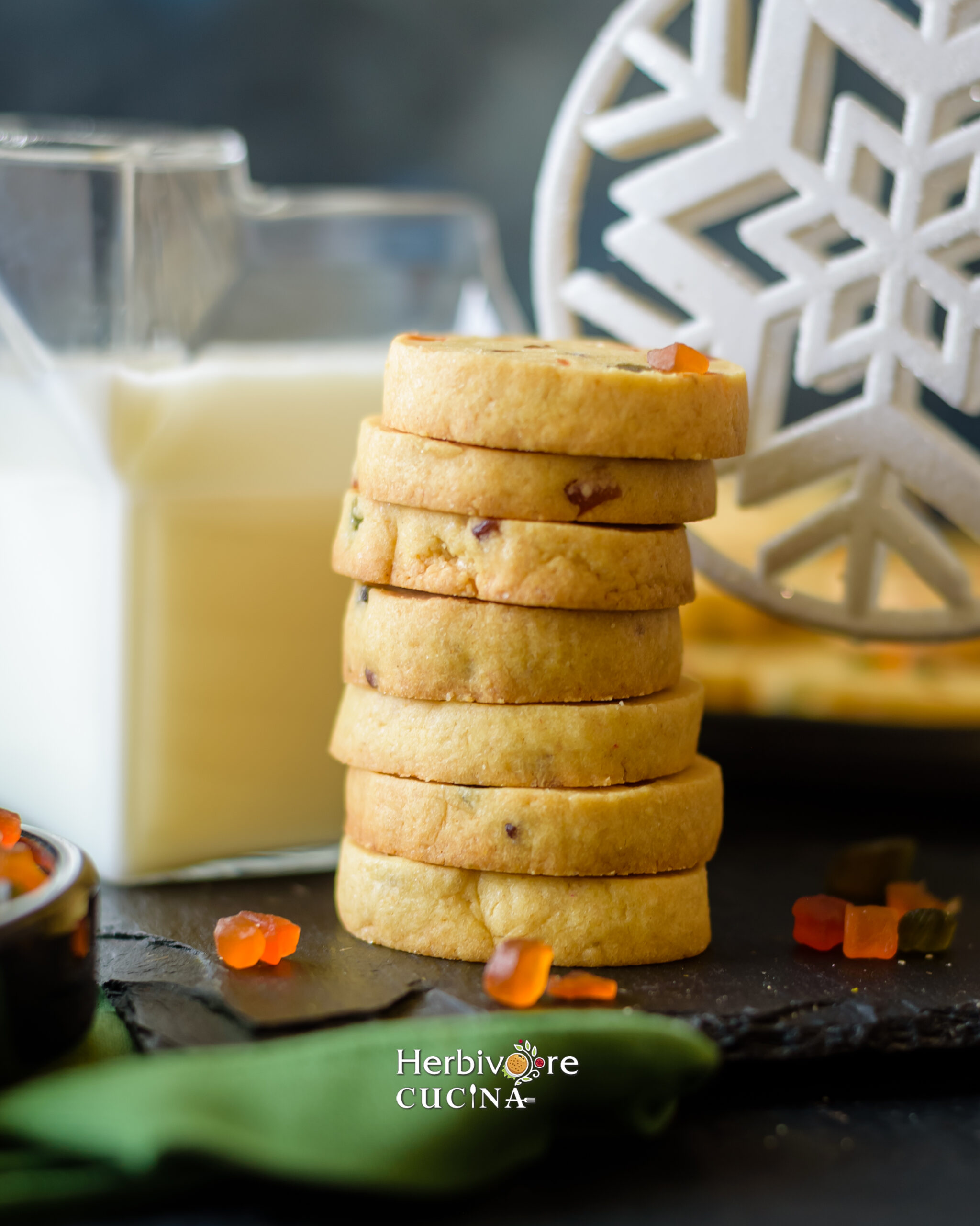 Piled eggless karachi cookies on a slate platter with a jar of milk behind it. 