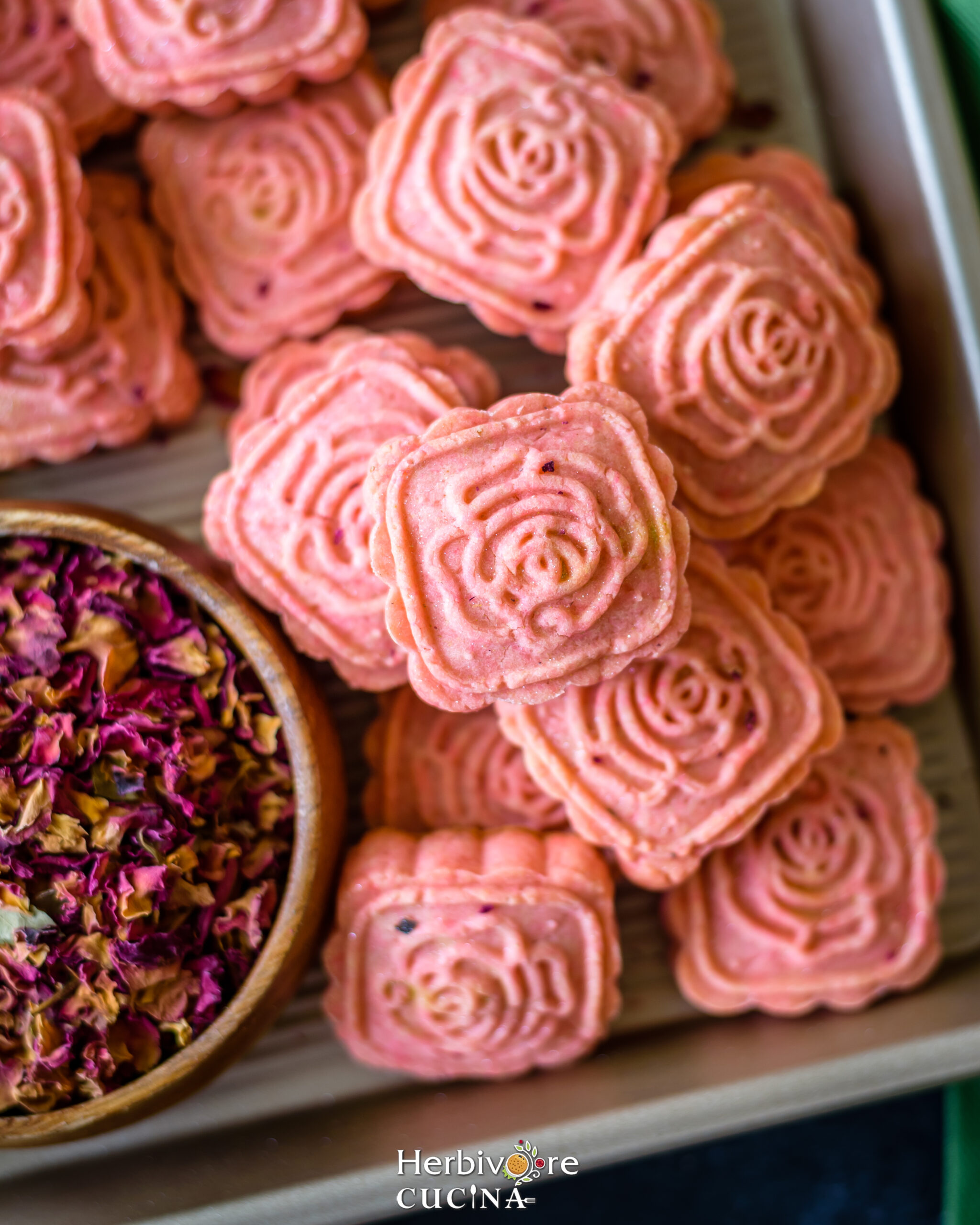 A baking tray with rose cardamom cookies and some dried rose petals in a small wooden bowl. 