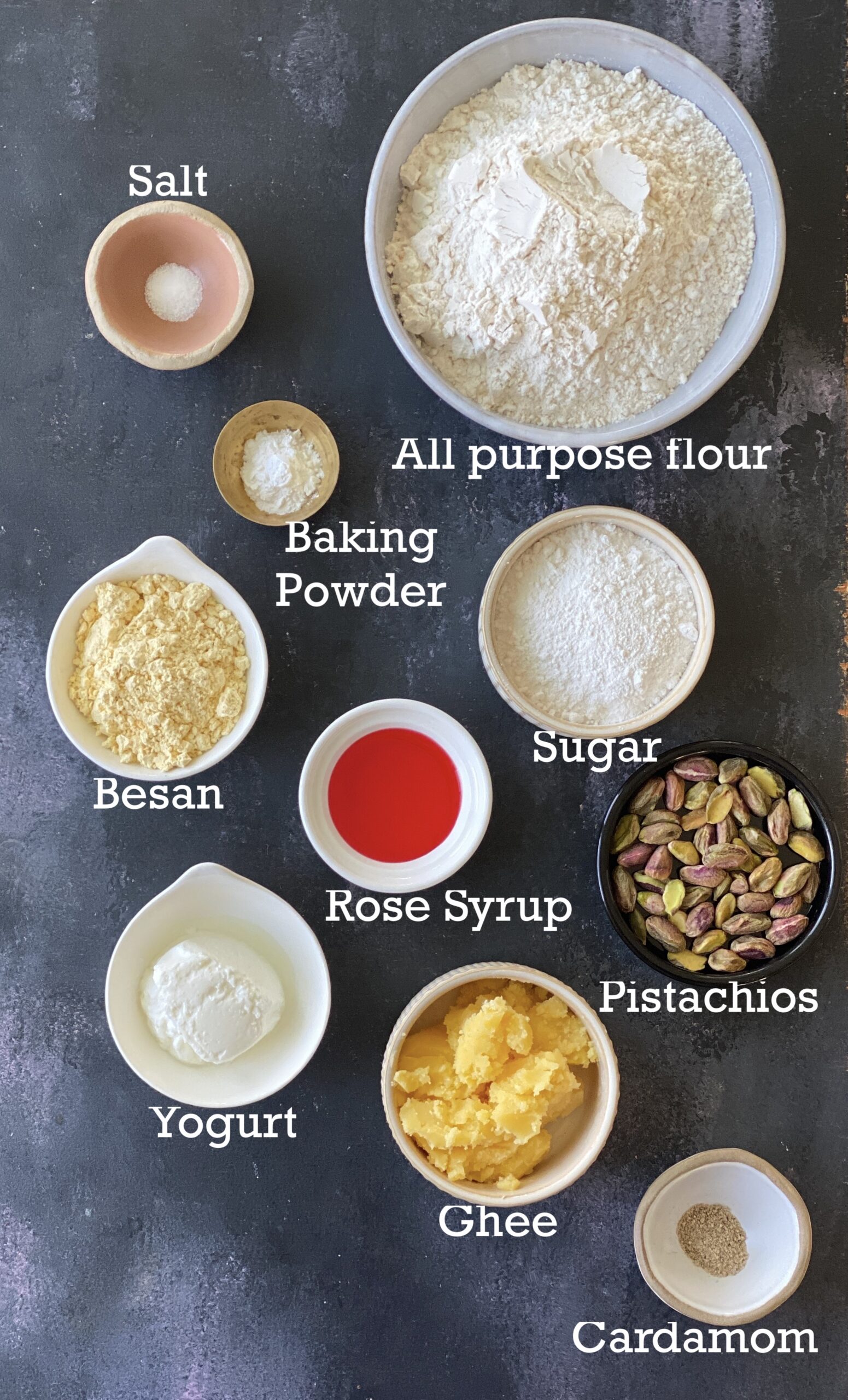 Ingredients to make rose cardamom cookies in small bowls on a black surface. 