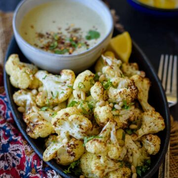 A black oval plate with air fryer tahini cauliflower with a tahini dip in gray bowl with a fork by the side and lemon in the background.
