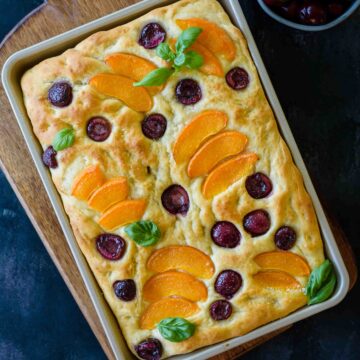 A tray with baked apricot and cherry focaccia ready to be sliced on a brown board.