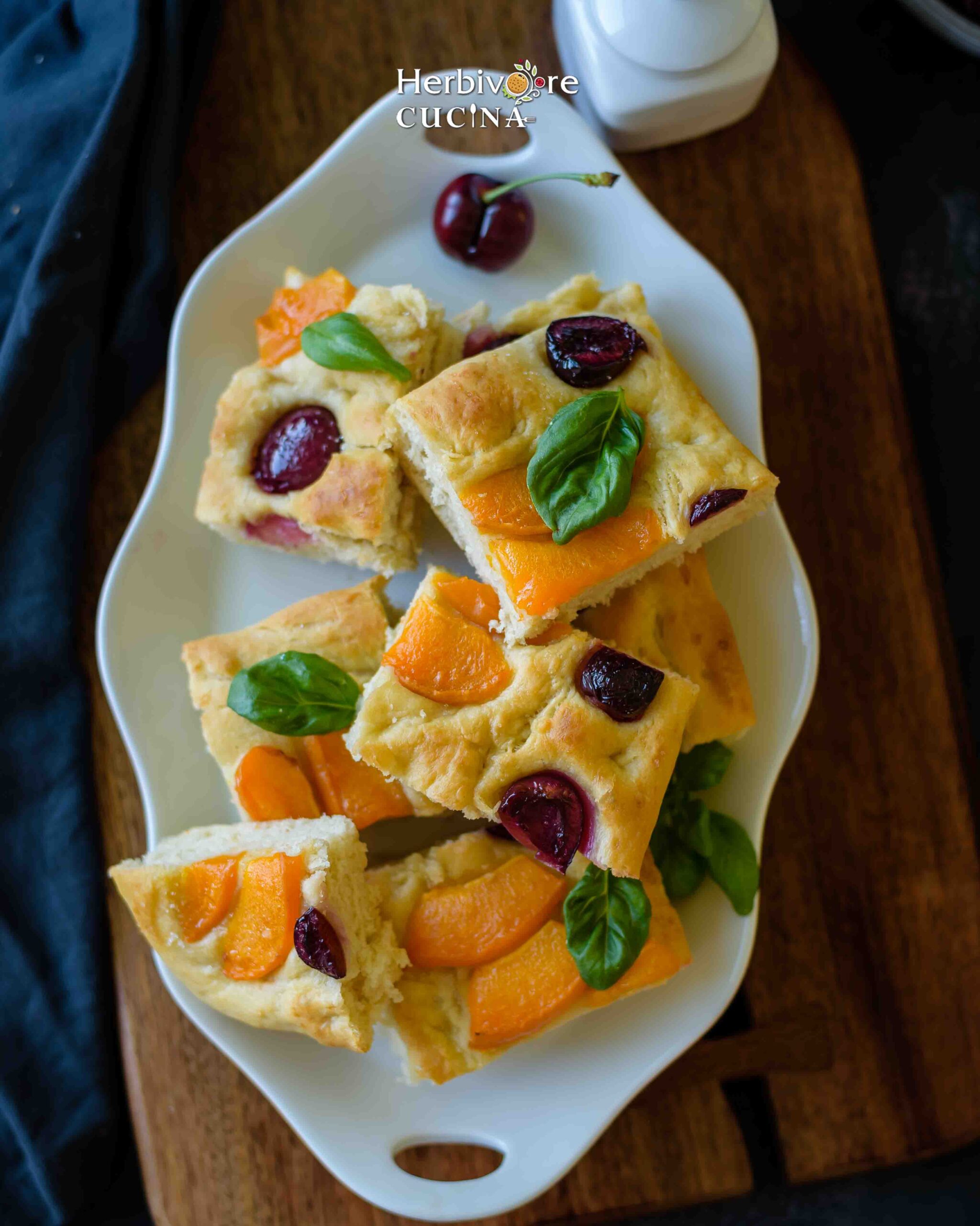 A white oval plate with slices of apricot and cherry focaccia topped with basil on a brown board. 