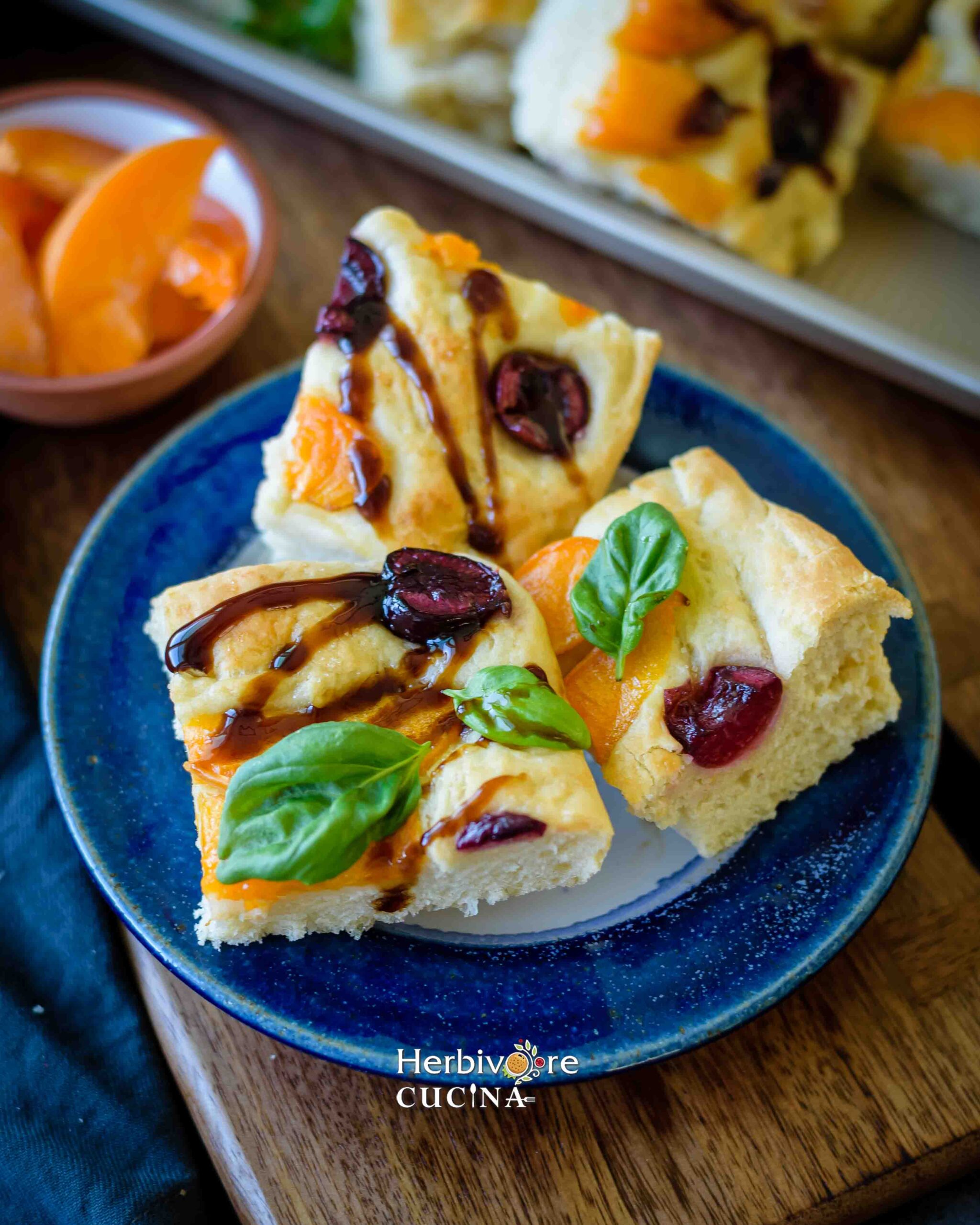 Serving apricot and cherry focaccia slices topped with balsamic vinegar with slices of apricot on the side on a brown board. 