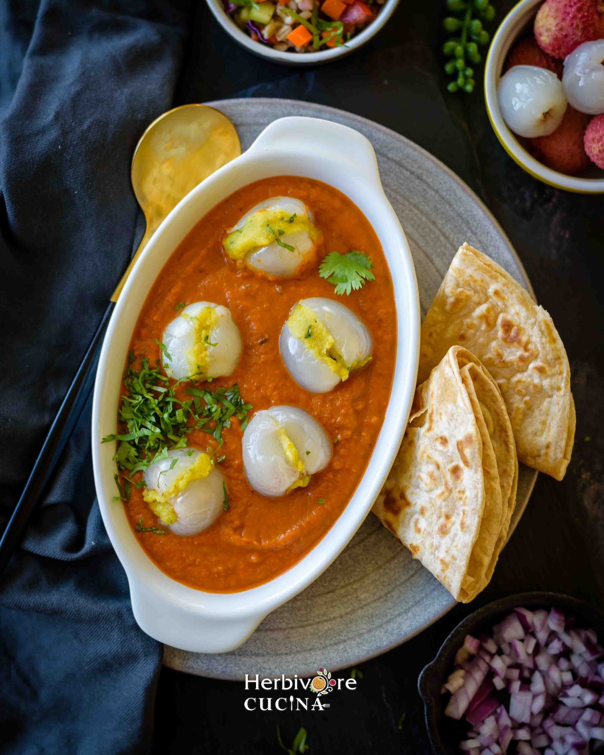 Litchi Kofta served in a white bowl with roti and salad on the side on a gray plate with a spoon and napkin around it. 