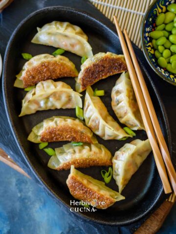Pan fried dumplings on a black oval plate with chopsticks on the side and some edamame placed around it.