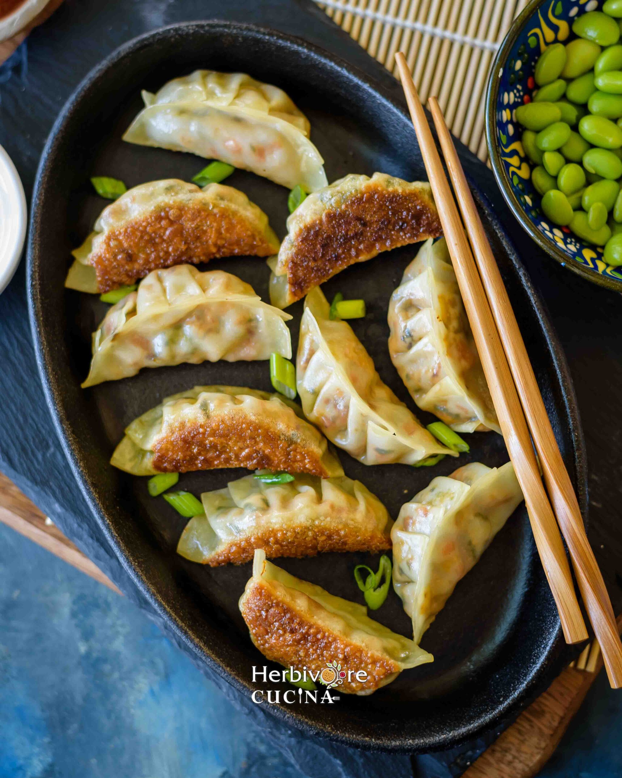 Pan fried dumplings on a black oval plate with chopsticks on the side and some edamame placed around it. 