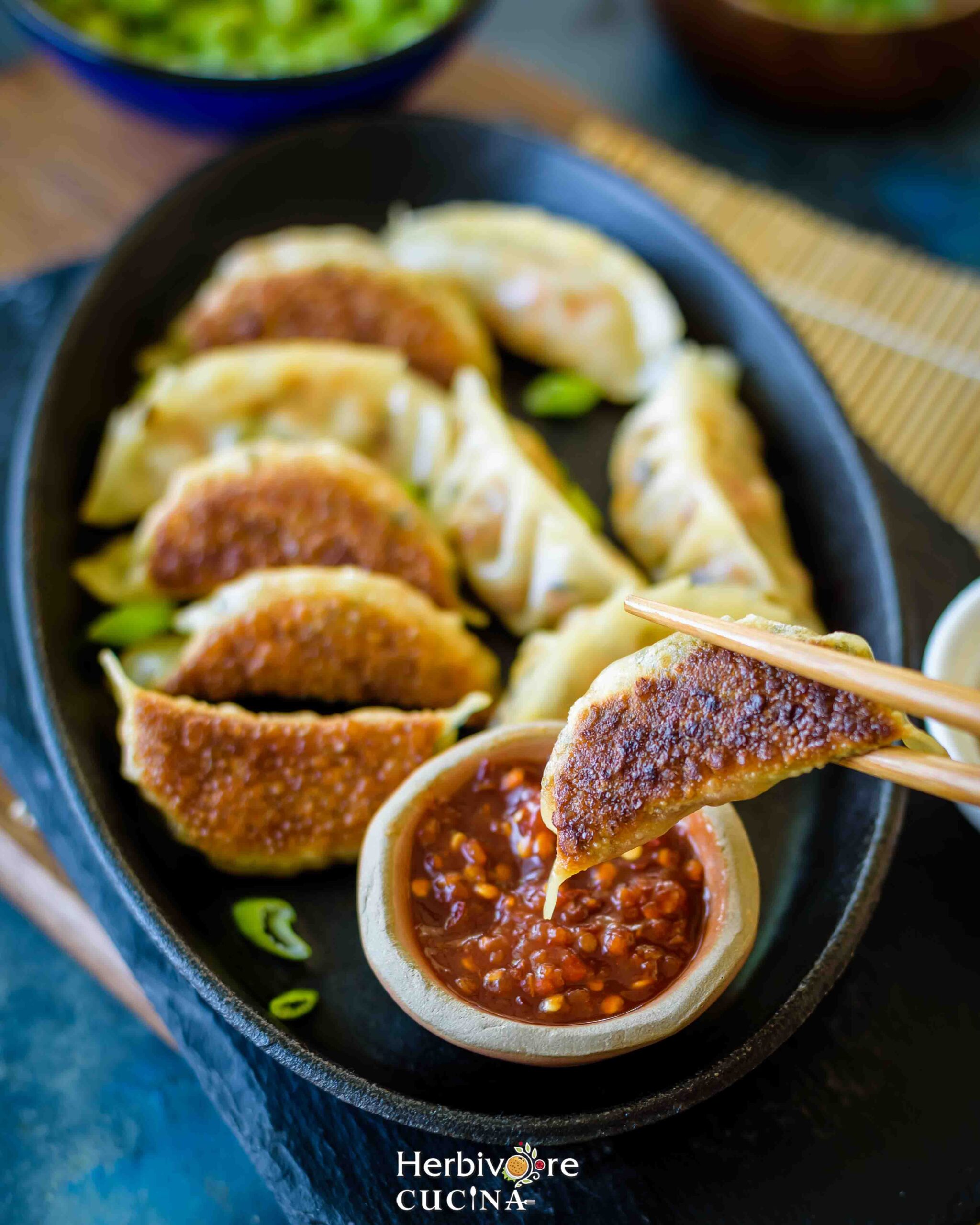 Black oval plate with hot sauce in a bowl; with a dumpling held in chopsticks above it. 