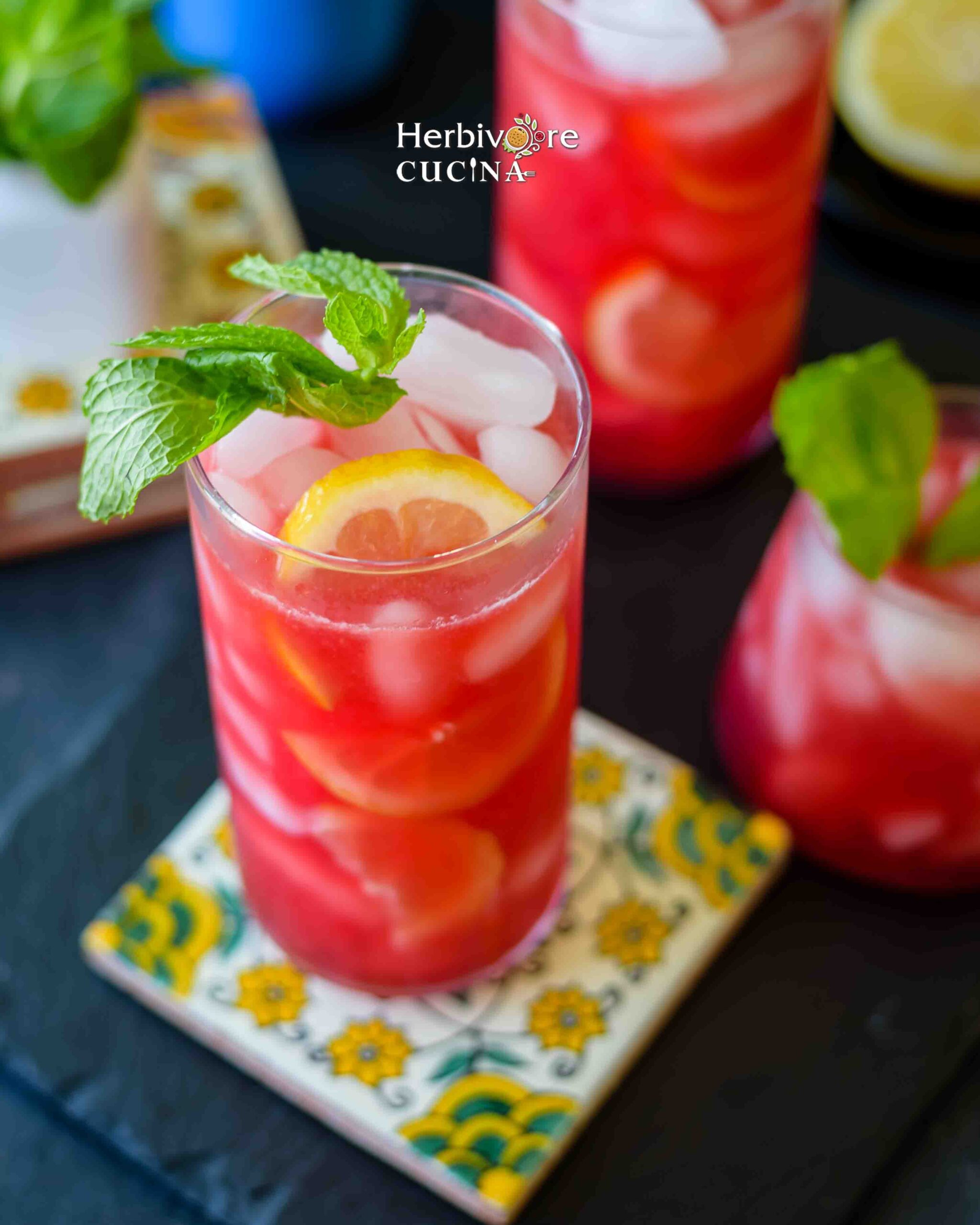 Side view of glass filled with ice and prickly pear lemonade topped with mint leaves and lemons on a colored coaster and slate platter.