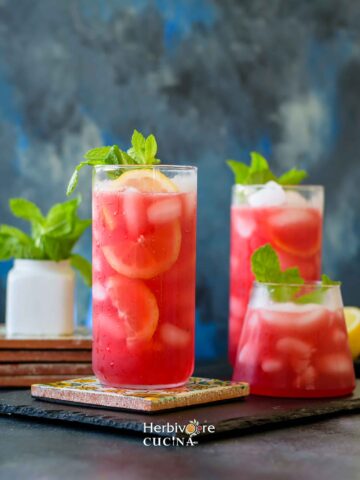 Three glasses filled with prickly pear lemonade topped with lemon, ice and mint with some mint leaves around it on a slate platter.