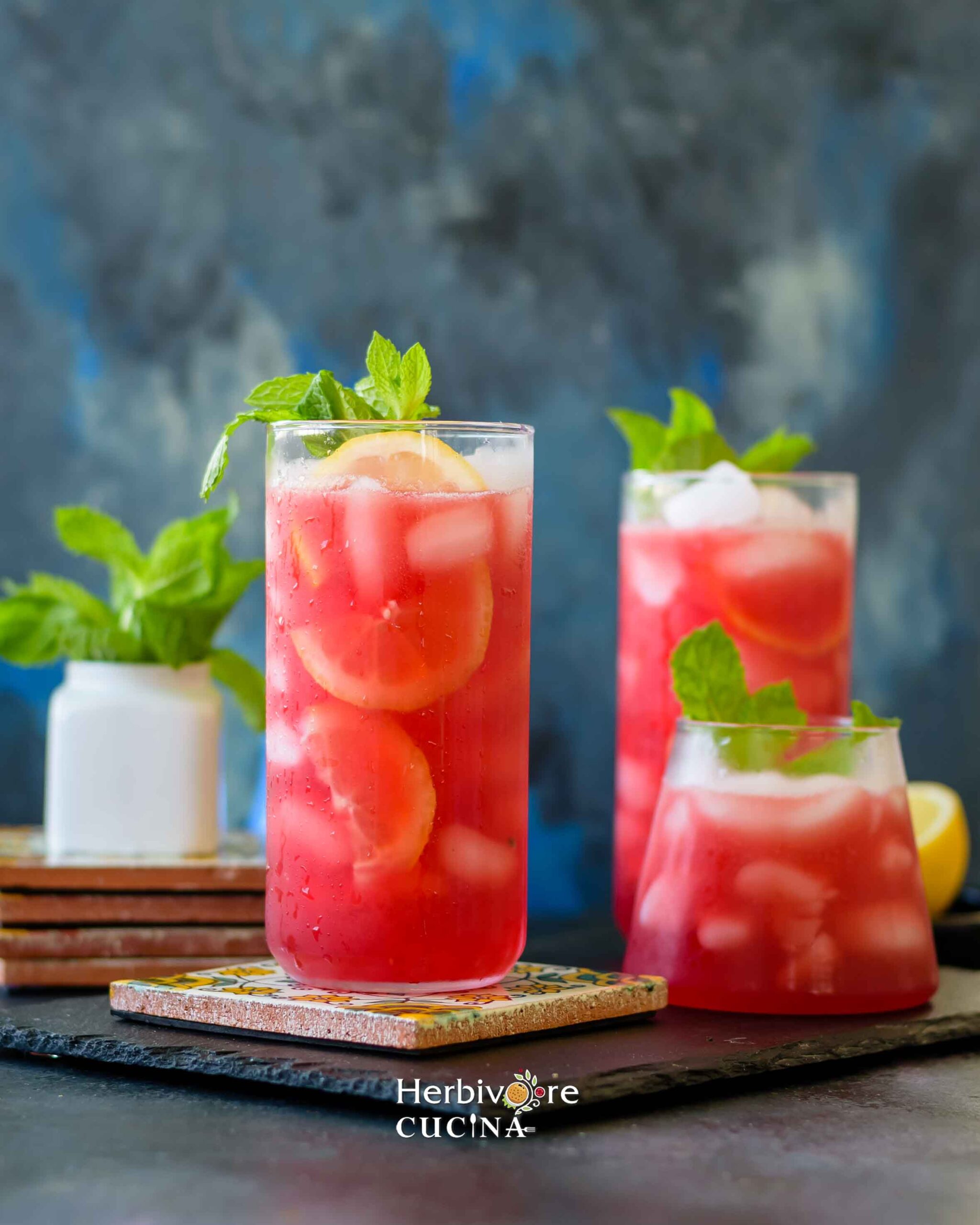 Three glasses filled with prickly pear lemonade topped with lemon, ice and mint with some mint leaves around it on a slate platter.