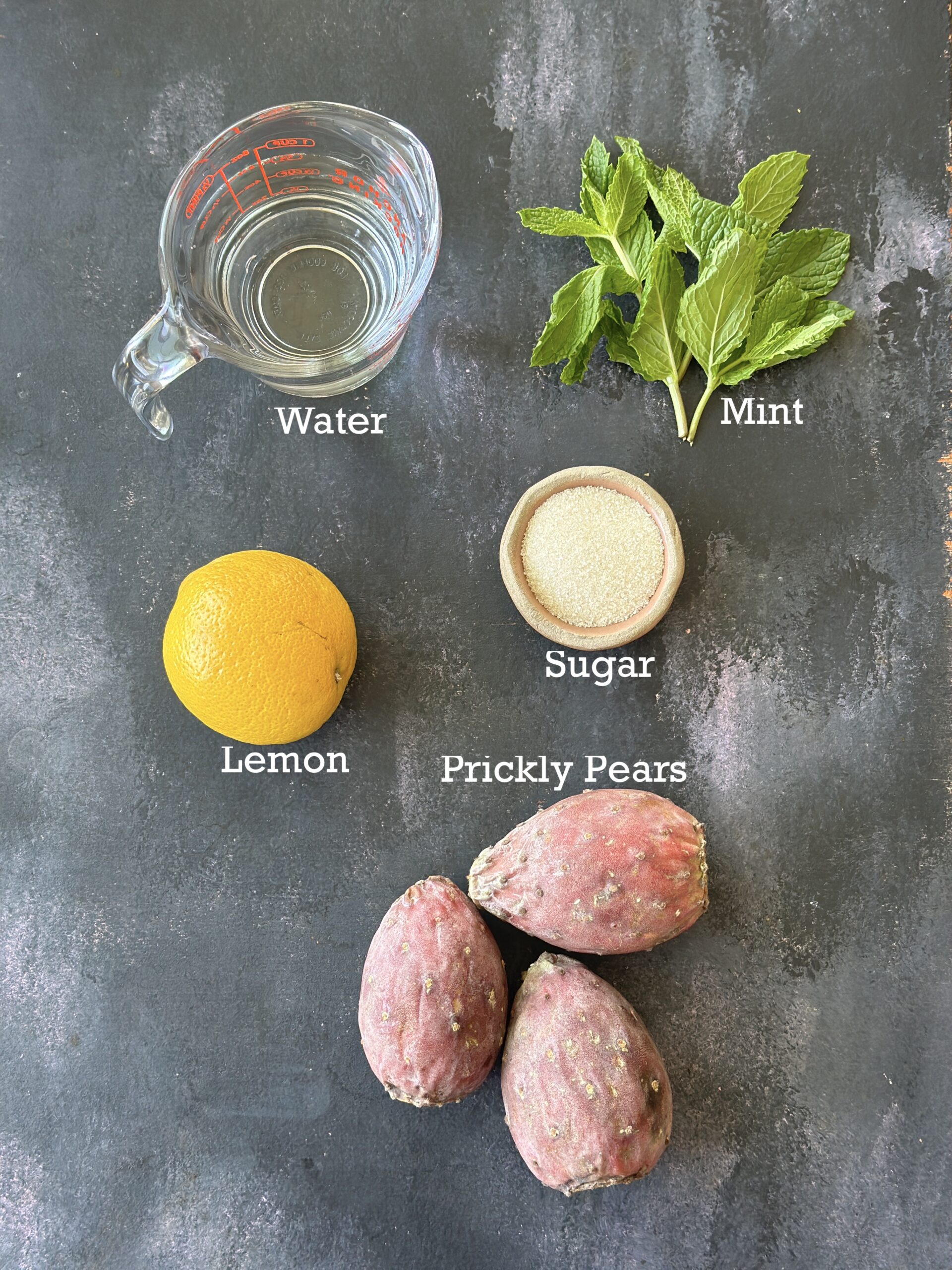 Ingredients for making Prickly pear Lemonade; pears, lemon, sugar, water and mint leaves on a black background.
