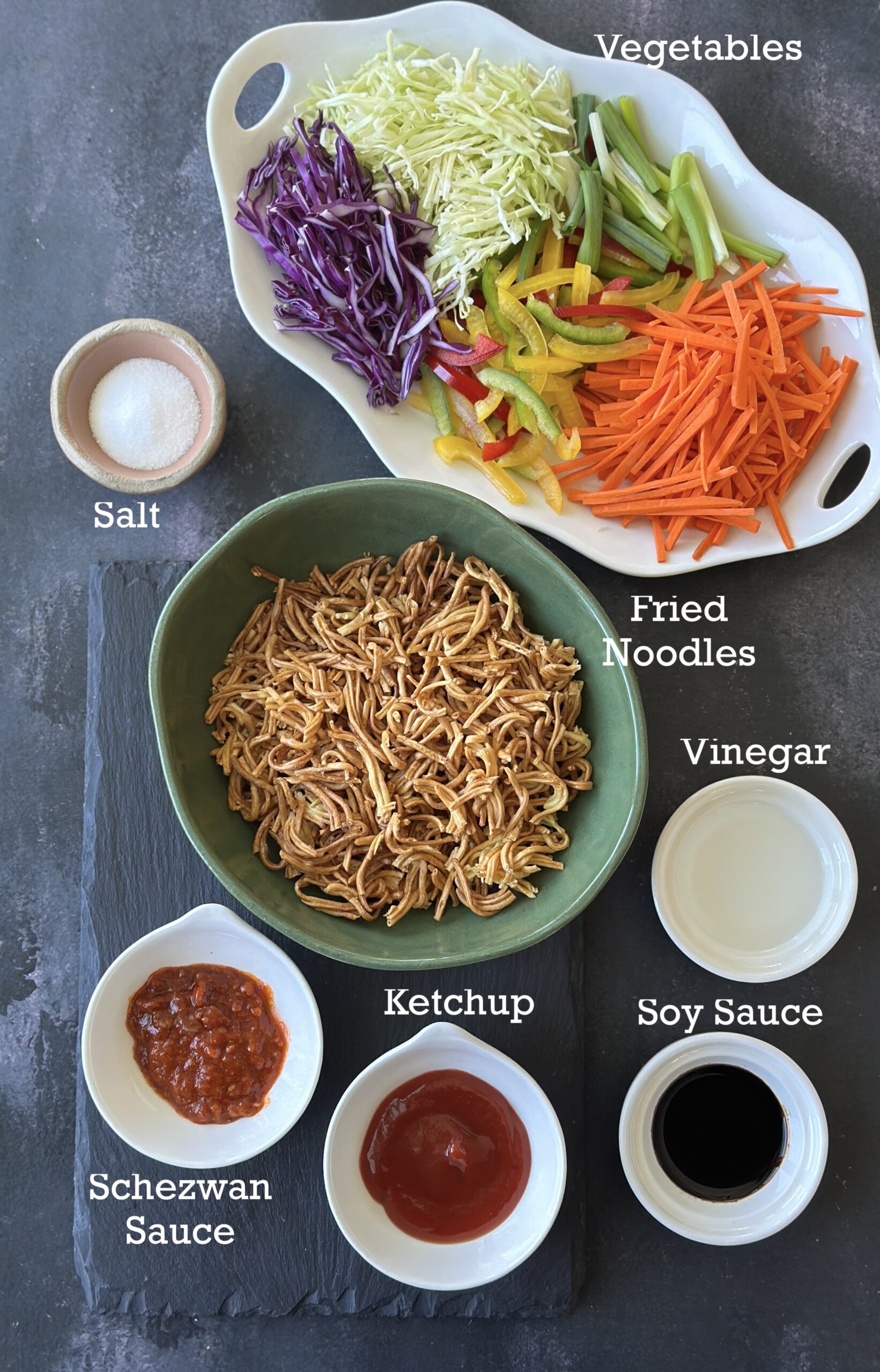 Ingredients to make Chinese Bhel; air fried noodles, vegetables, sauces and salt on a dark background. 