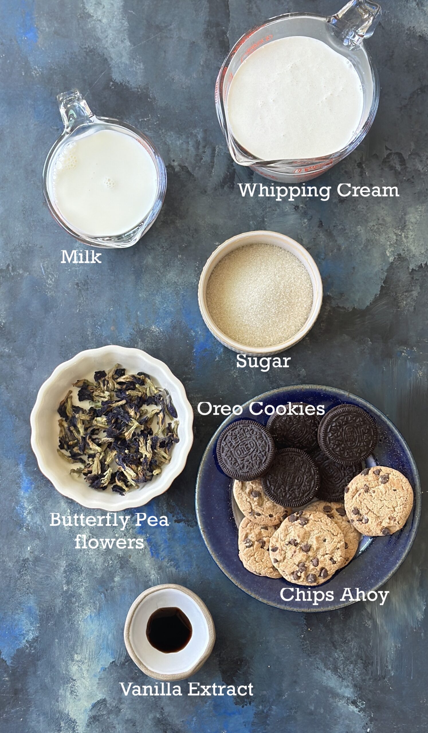 Ingredients for Cookie Monster ice cream; cream, milk, butterfly pea flowers and cookies on a blue black board. 