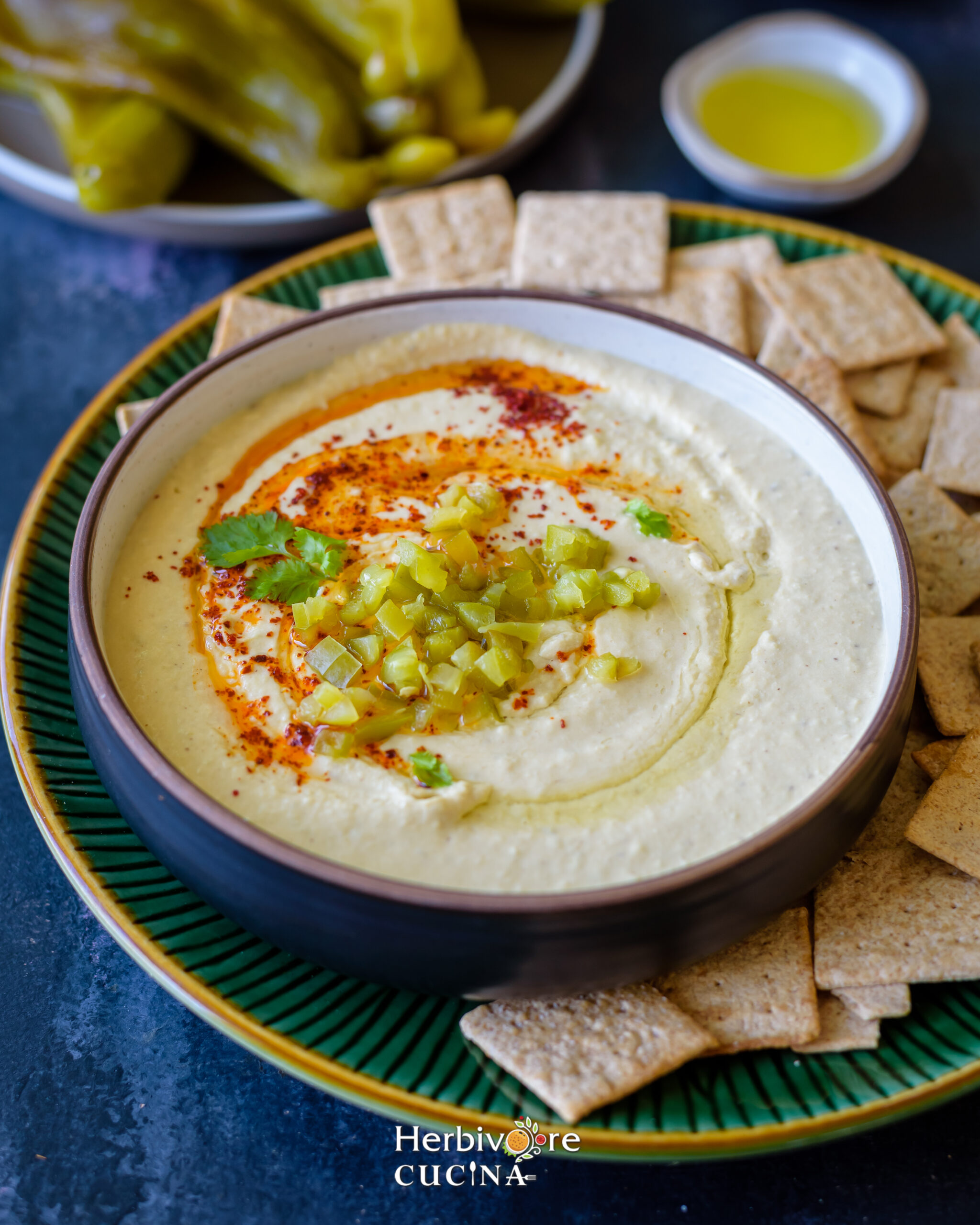 Side view of hatch chile hummus in a bowl with crackers on the side and more chilis in the background. 