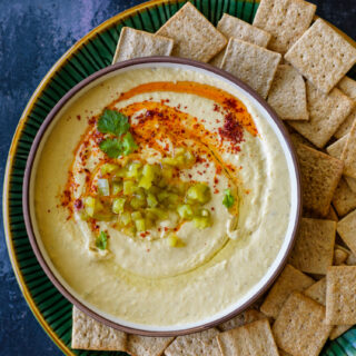 A brown bowl filled with Hatch Chile Hummus topped with oil and paprika and served with crackers in a green plate.