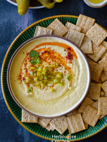 A brown bowl filled with Hatch Chile Hummus topped with oil and paprika and served with crackers in a green plate.